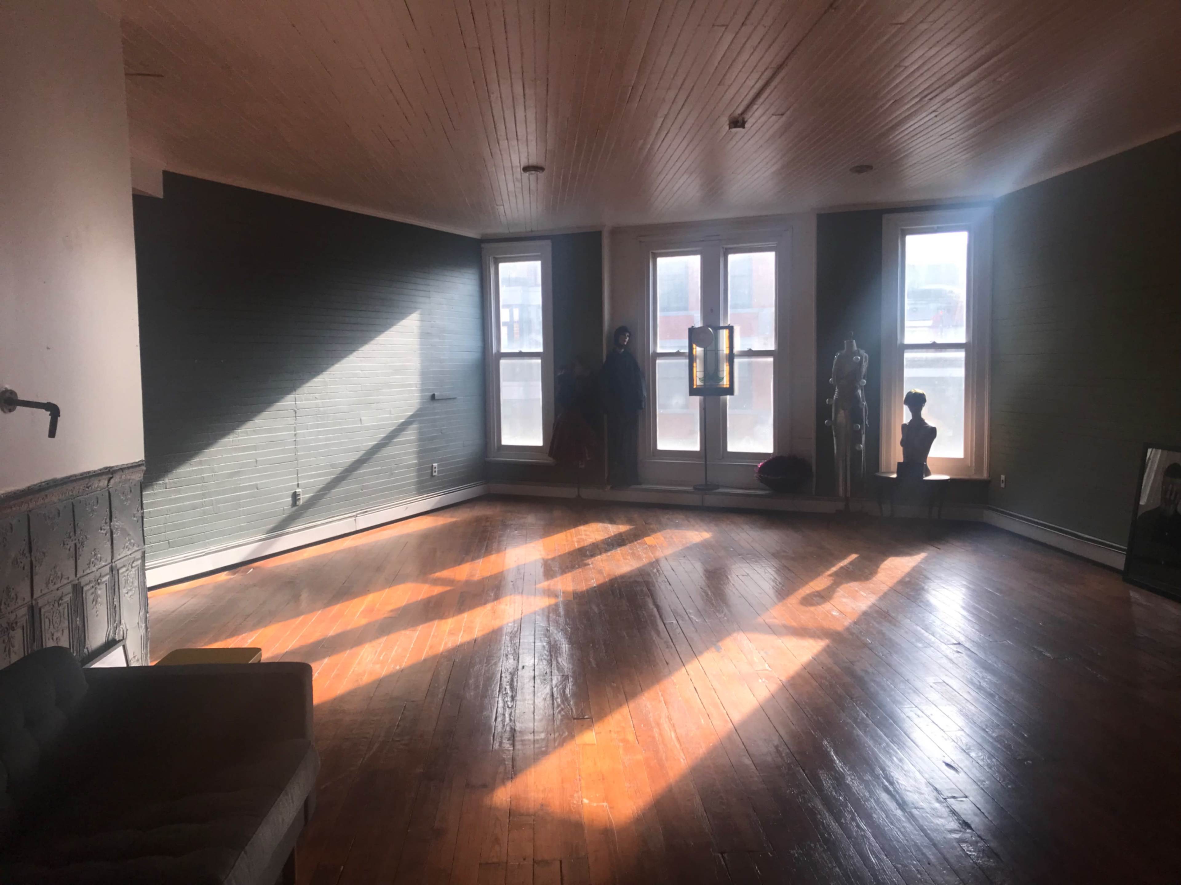 The image shows a spacious room with hardwood floors, large windows allowing sunlight to stream in, and a few decorative elements including a sofa and mannequins.