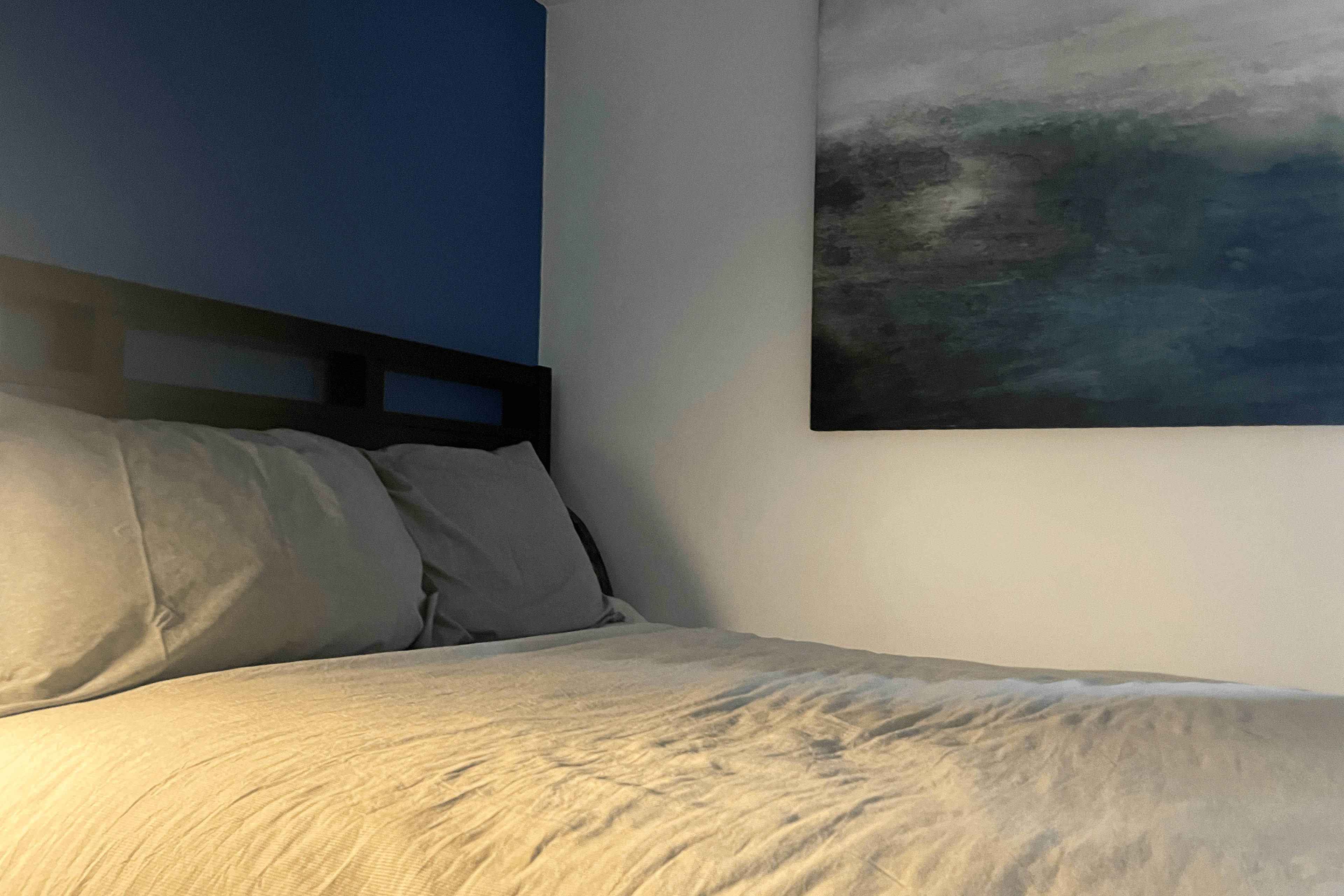 A neatly made bed with gray bedding against a wall painted blue, with a large abstract painting on the adjacent white wall.