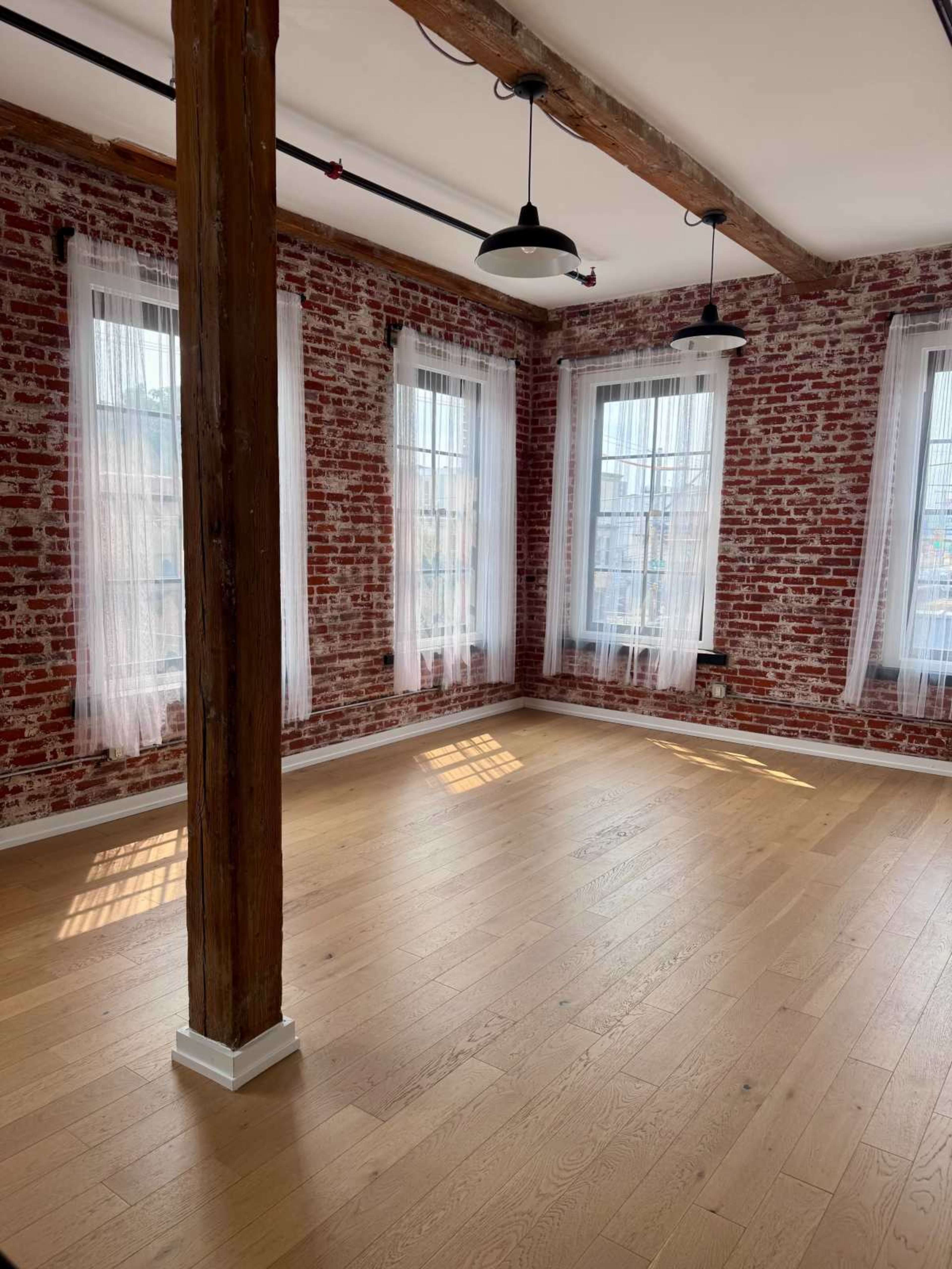 The image shows a spacious room with exposed brick walls, large windows dressed in sheer white curtains, and wooden beams overhead.