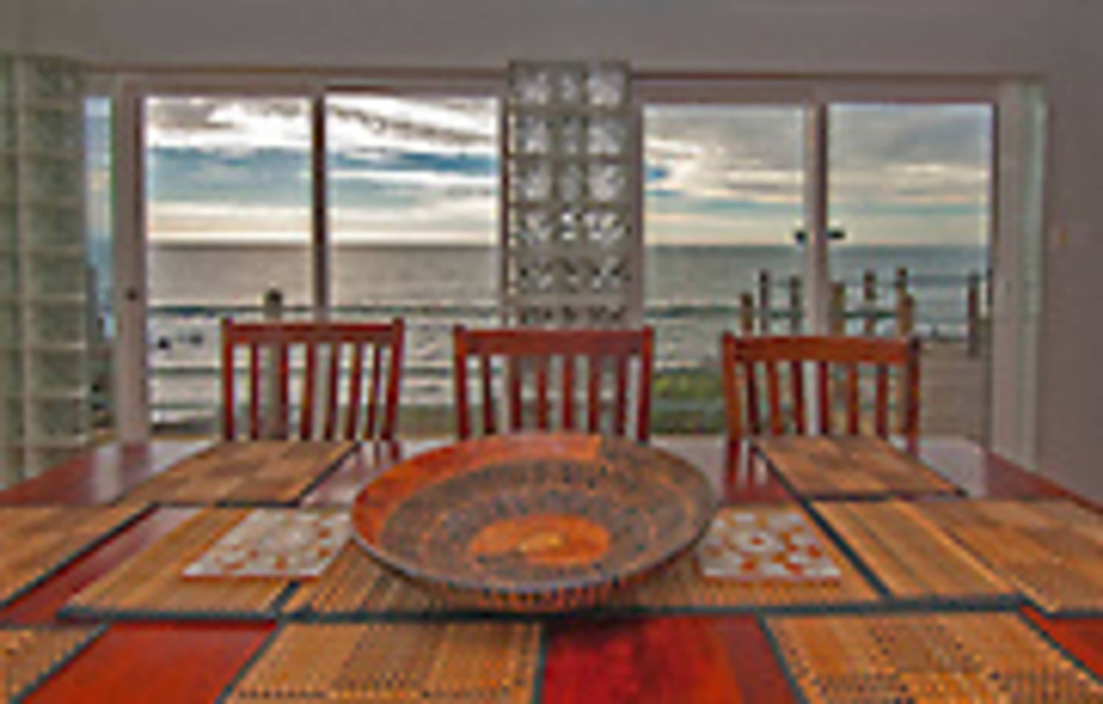 A dining table with a decorative bowl is set against a backdrop of a shoreline view visible through large glass windows.
