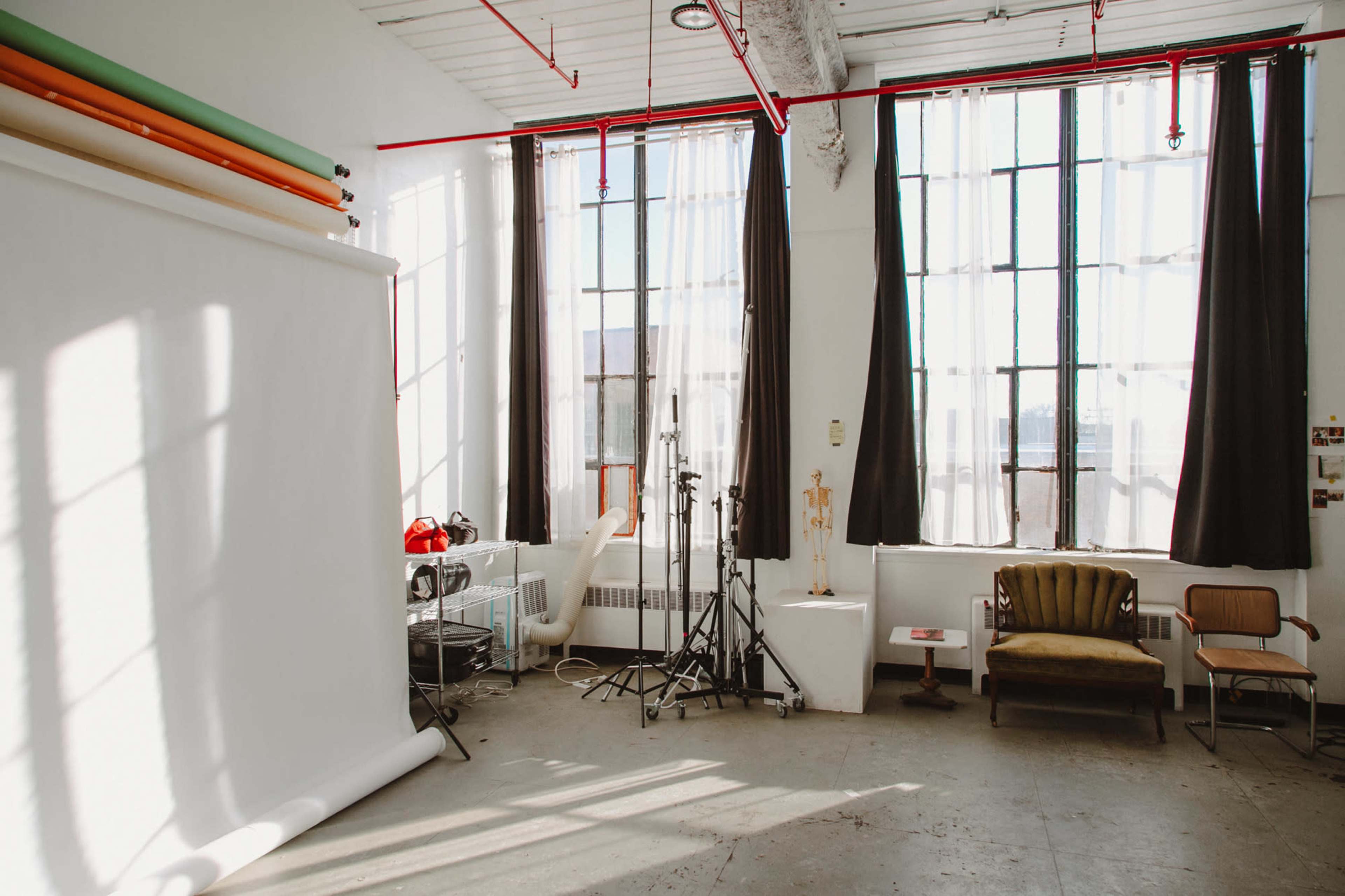 The image shows a bright studio space with large windows, a white backdrop, and simple furniture including a sofa and chair.
