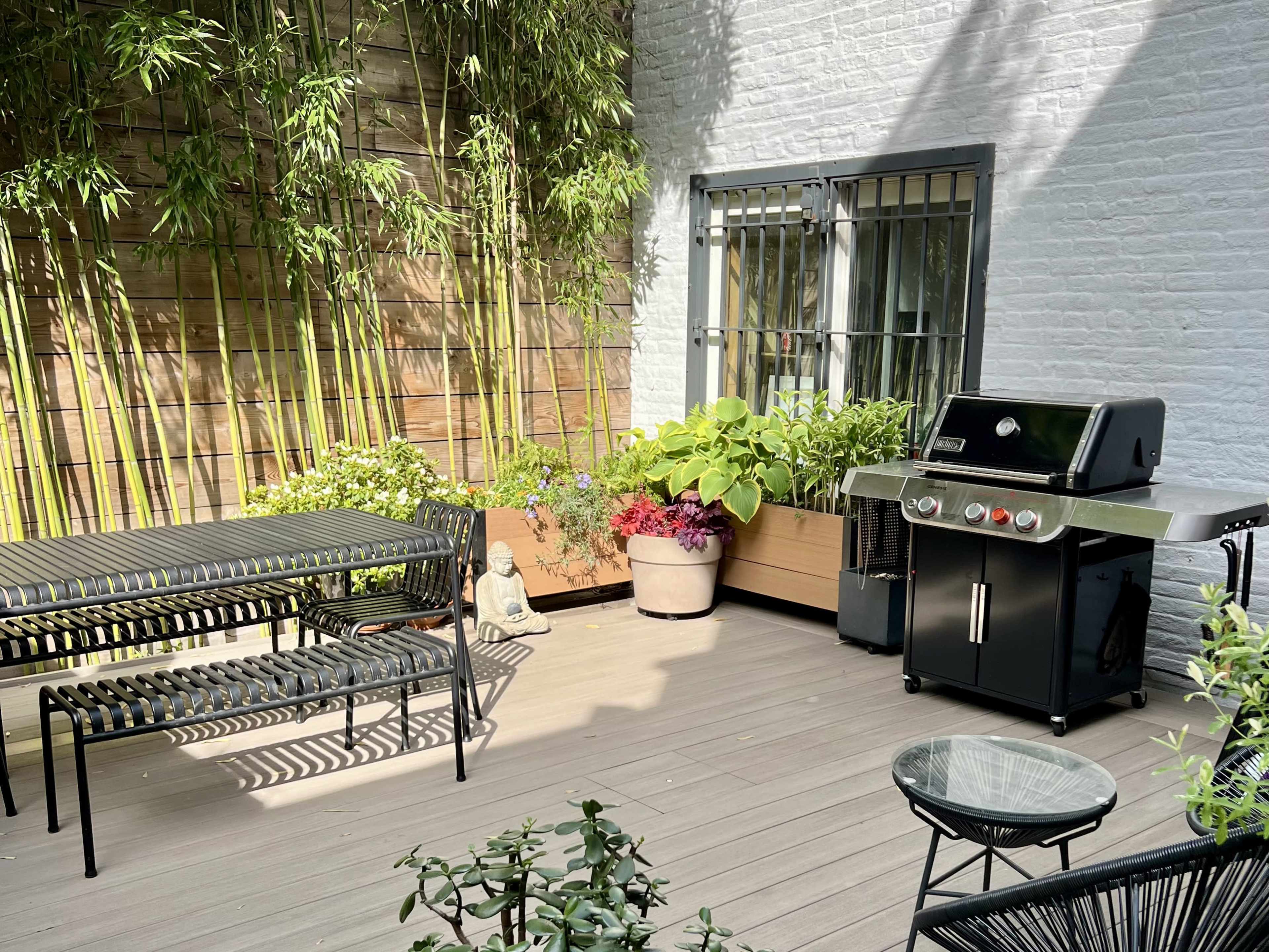 The image depicts a patio with a black grill, a glass table, and several metal chairs surrounded by potted plants and bamboo.