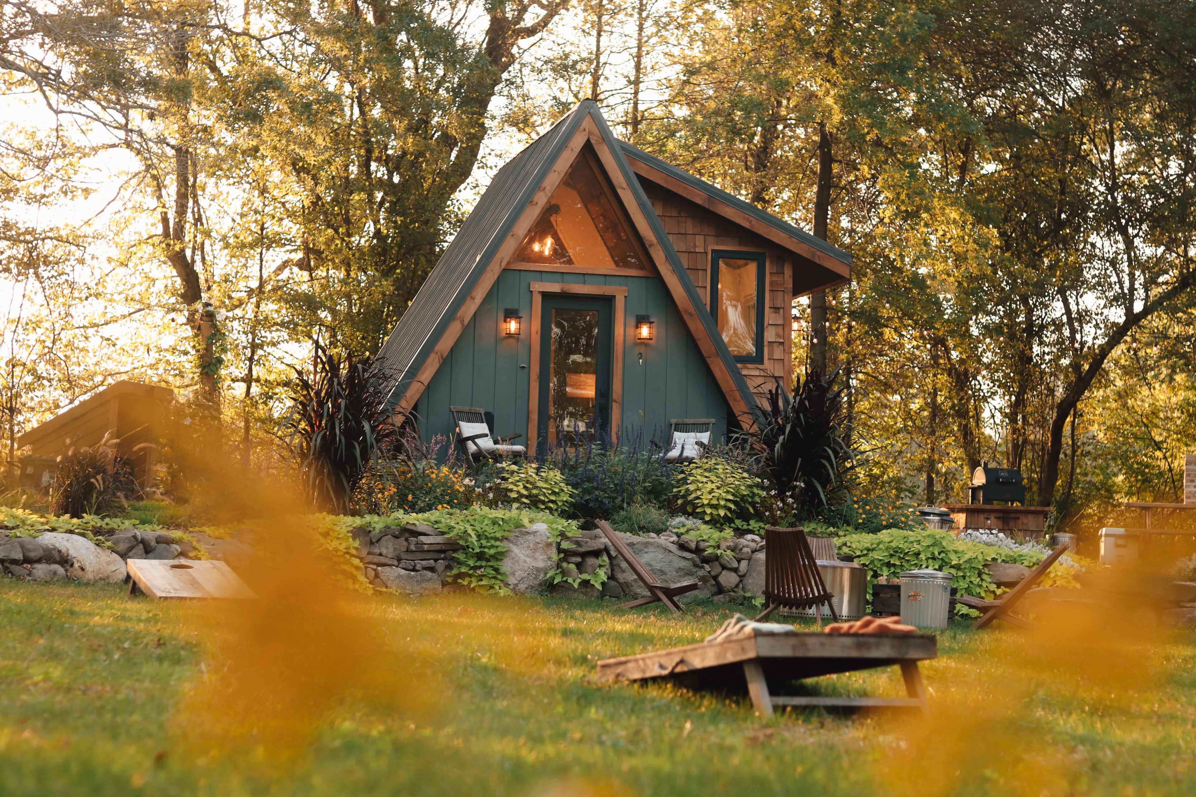 A small A-frame cabin with green siding is surrounded by trees and a grassy yard featuring lounge chairs and a stone border.