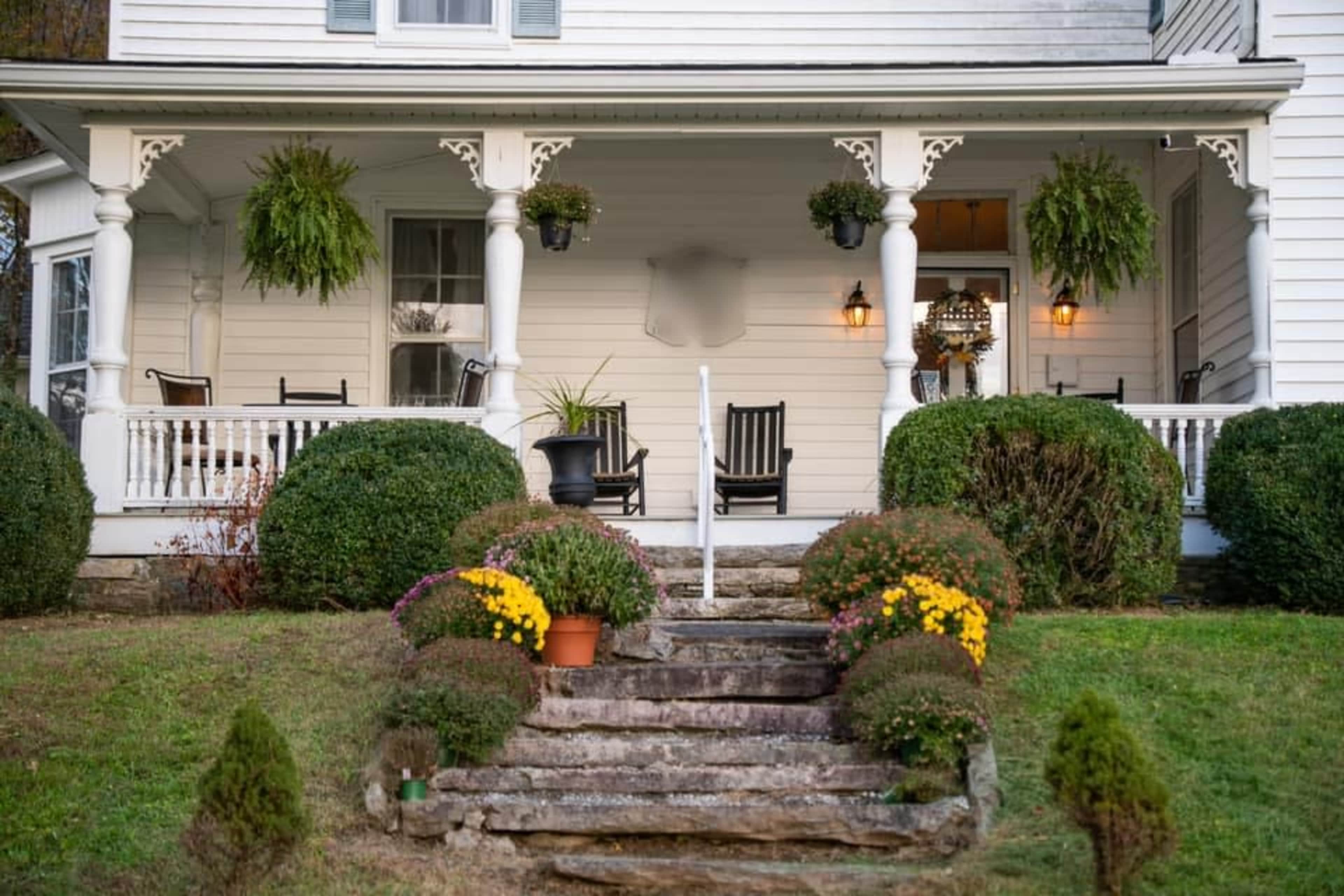 A porch of a white house with black rocking chairs, hanging plants, and flower pots, featuring stone steps leading up to it.
