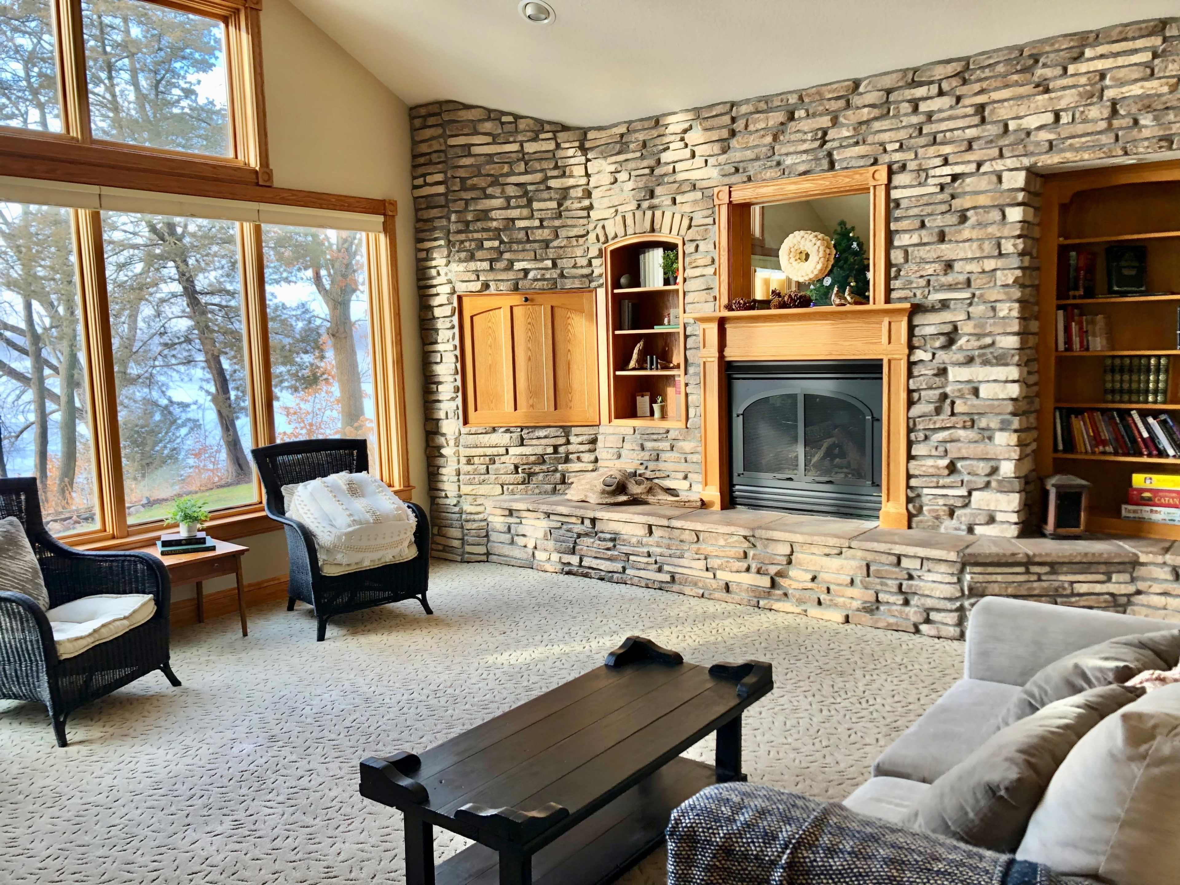 The image depicts a cozy living room featuring a stone fireplace, large windows allowing natural light, and rustic furnishings.