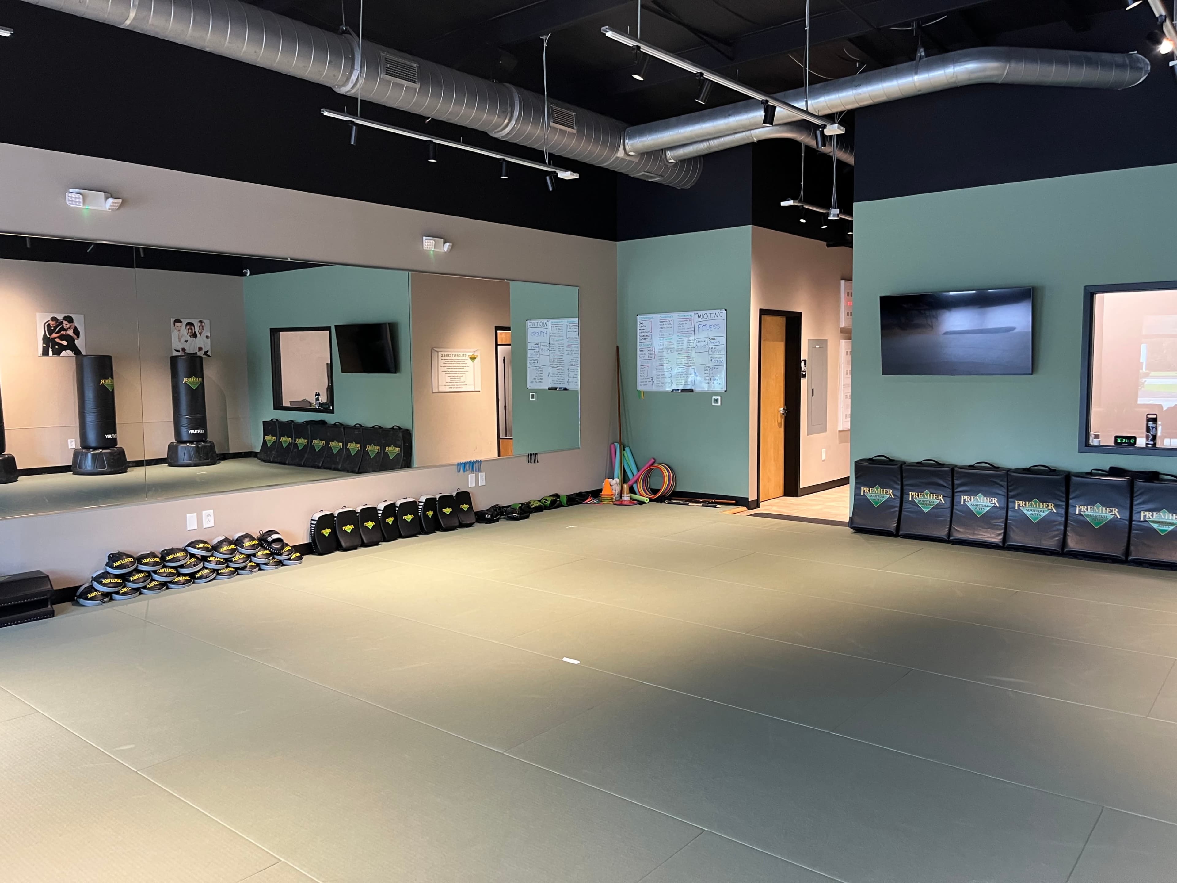 The image shows a fitness studio with a padded floor, mirrored walls, wall-mounted television, and various workout equipment neatly arranged along the sides.