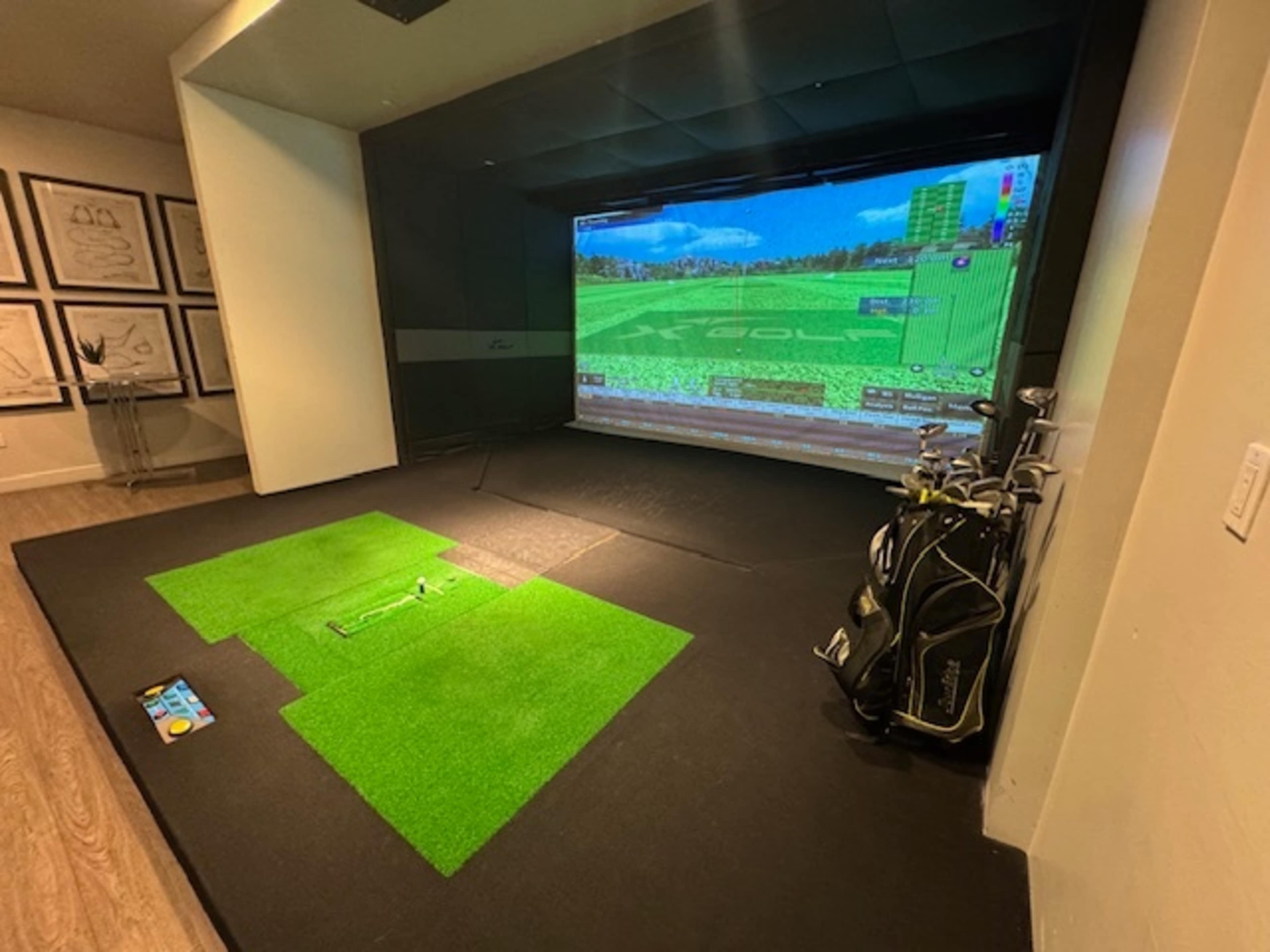 The image shows an indoor golf simulator setup with artificial turf, a large projection screen displaying a golf course, and golf clubs arranged nearby.