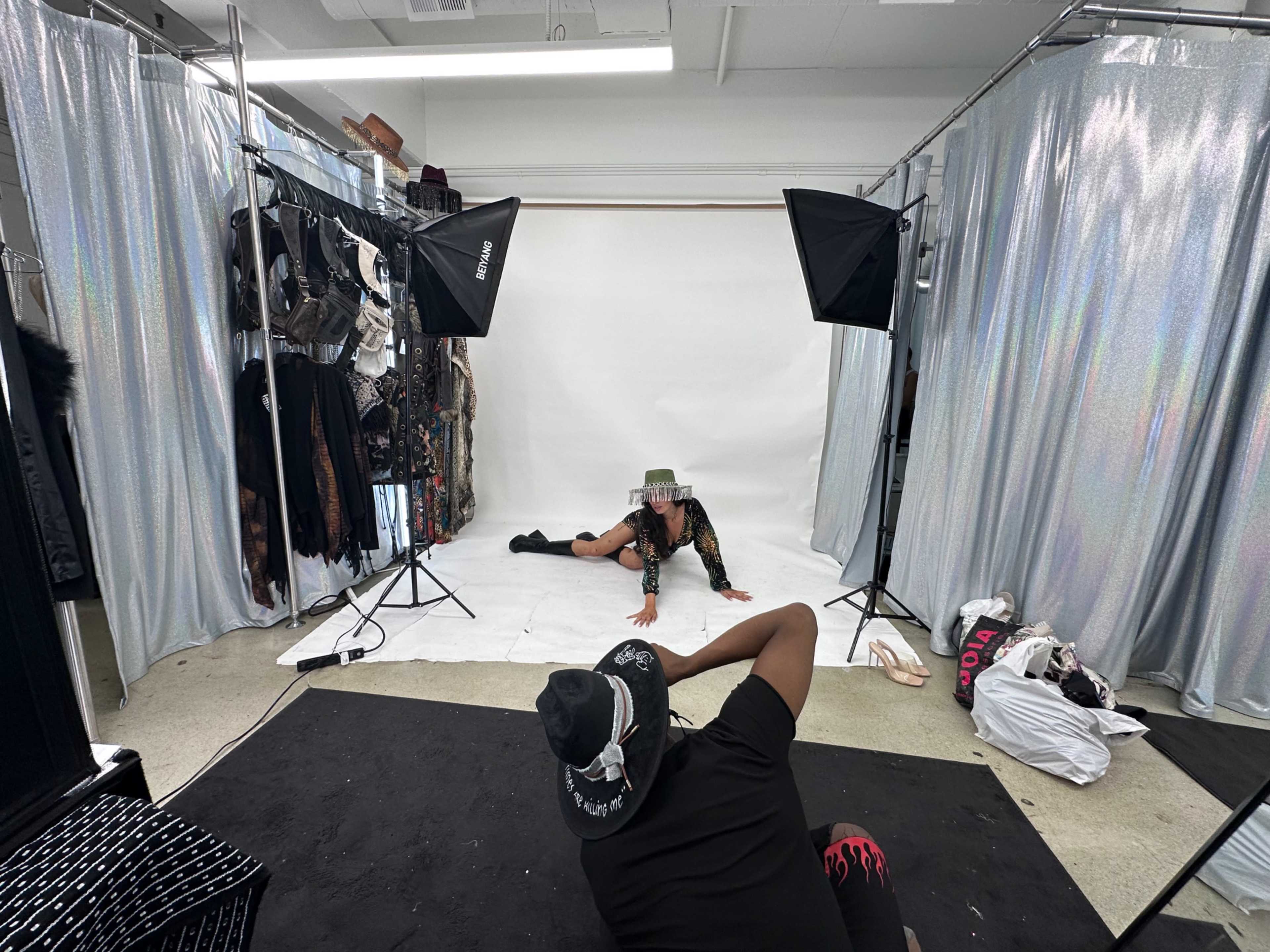A model poses on the ground in a fashion studio surrounded by clothing racks and photography equipment.