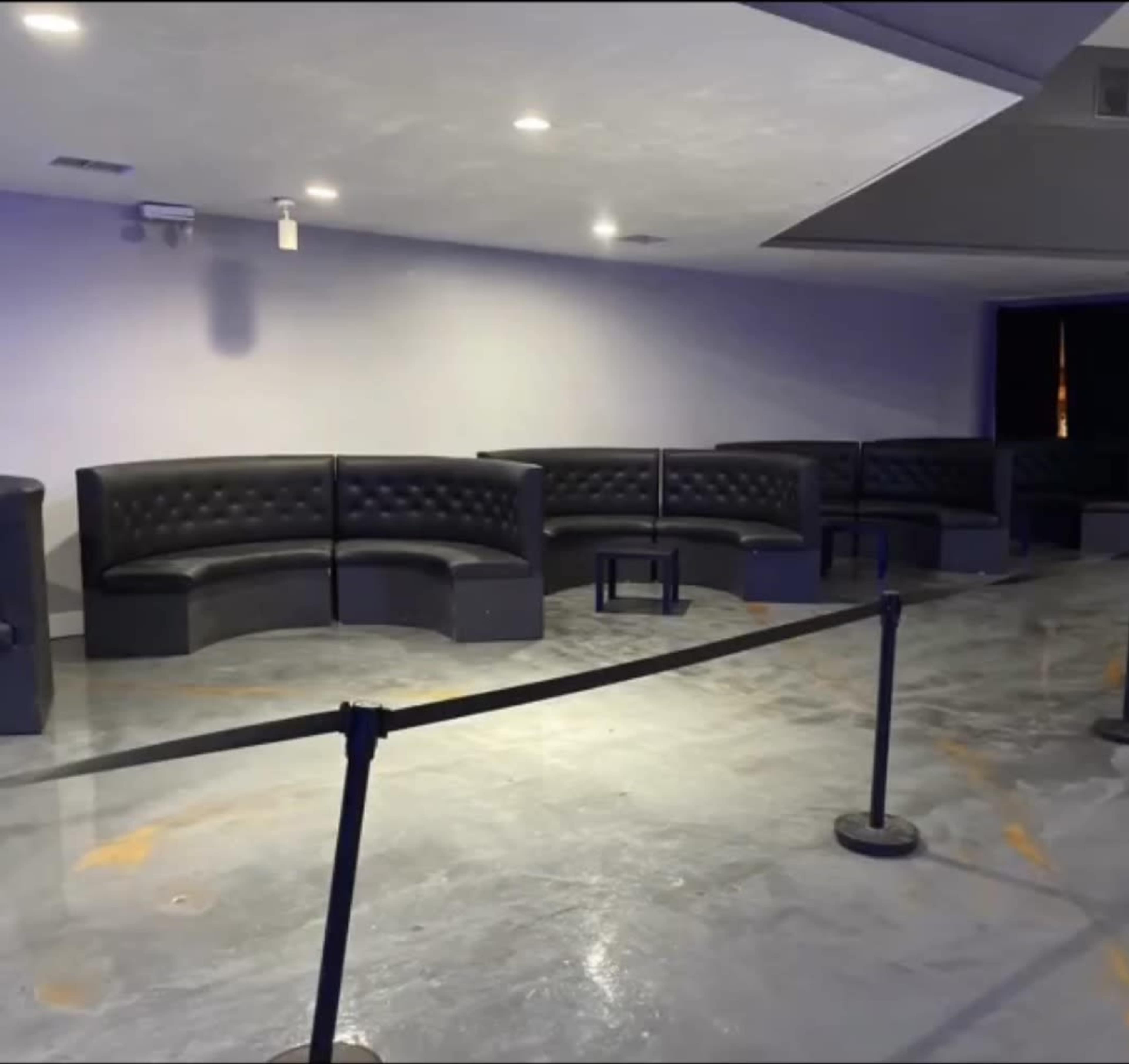 The image shows a dimly lit lounge area with black curved couches and a small table, separated by stanchions and ropes.