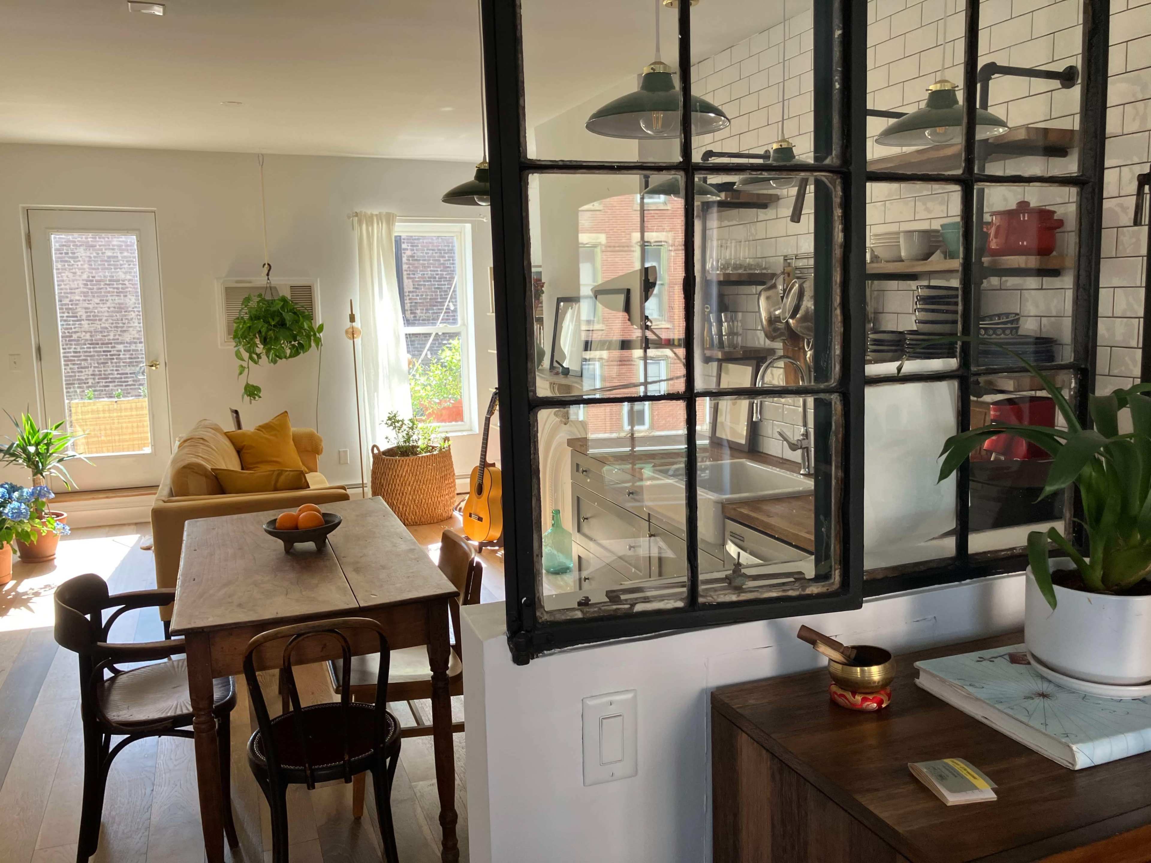 A cozy interior scene features a kitchen with glass partitions, a dining table with wooden chairs, and a bright living area with a yellow couch and plants.