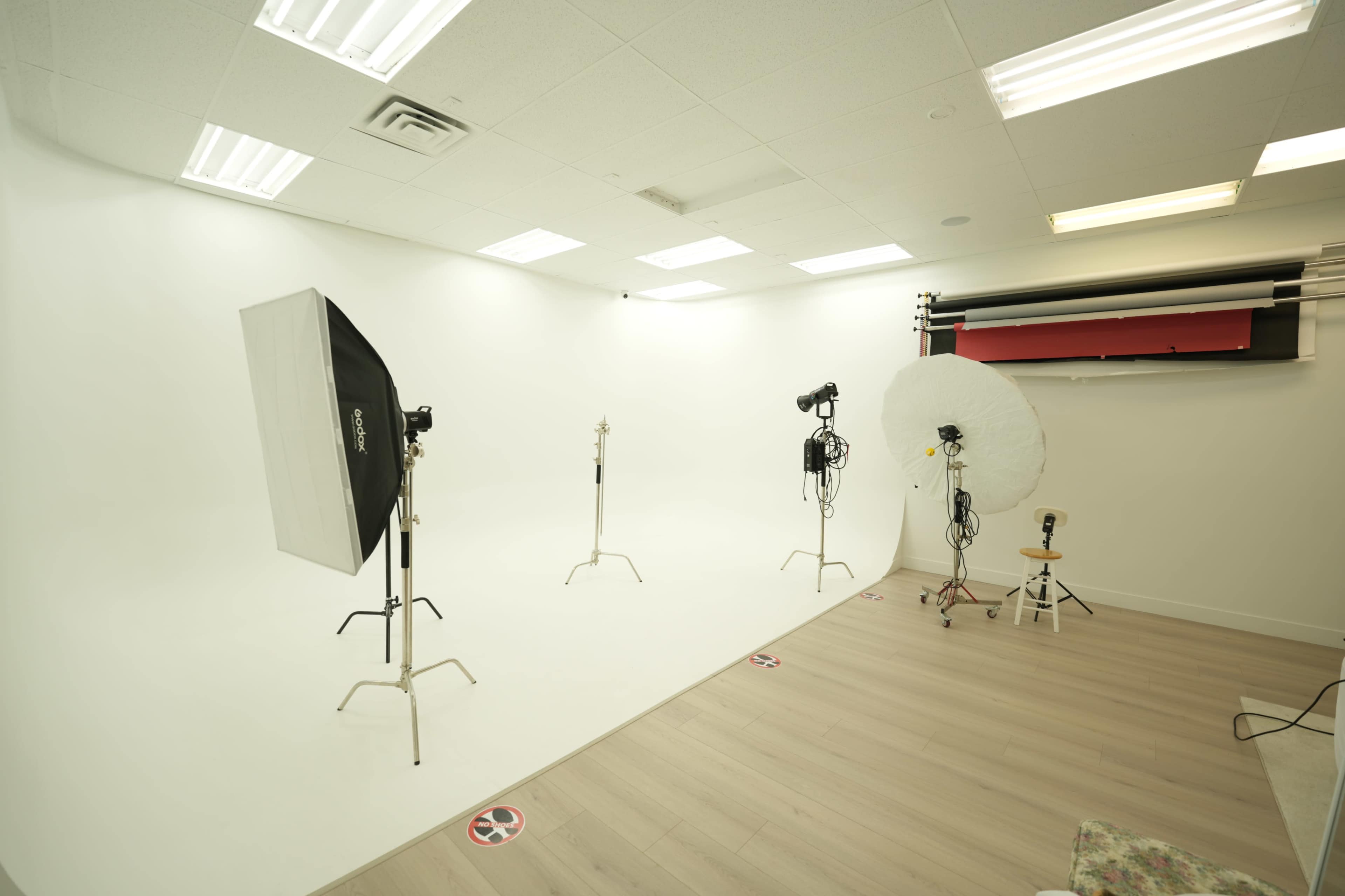 The image shows a bright, empty photography studio featuring a seamless white backdrop, two light stands with softboxes, and a round reflective umbrella.