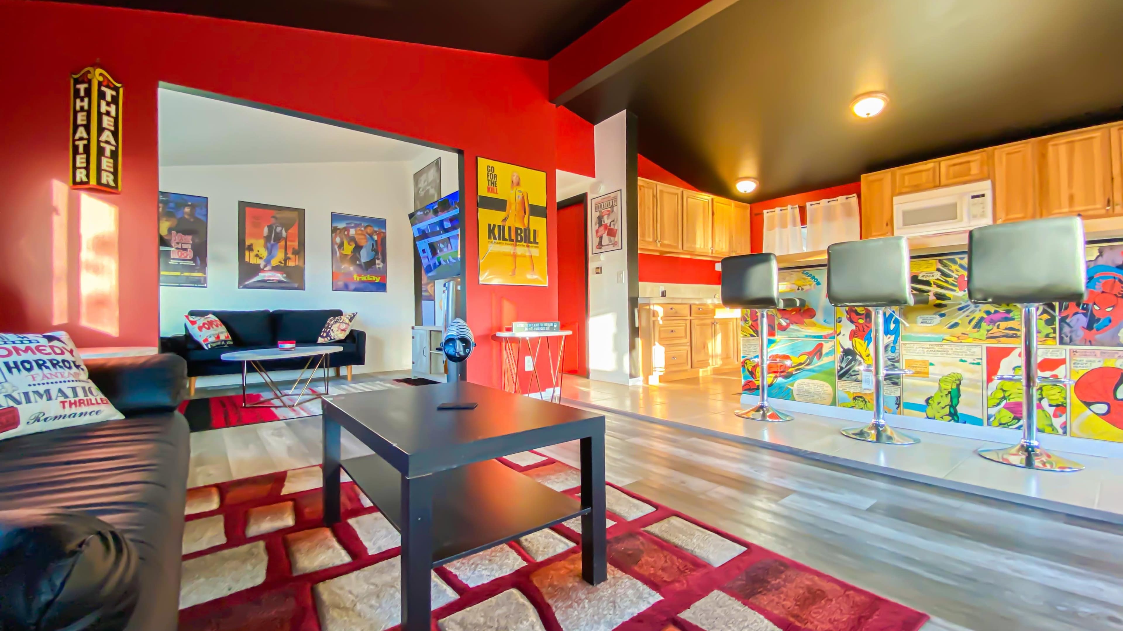 The image shows a modern, open-concept living space featuring a black leather sofa, colorful wall art, a kitchen area with bar stools, and hardwood flooring.