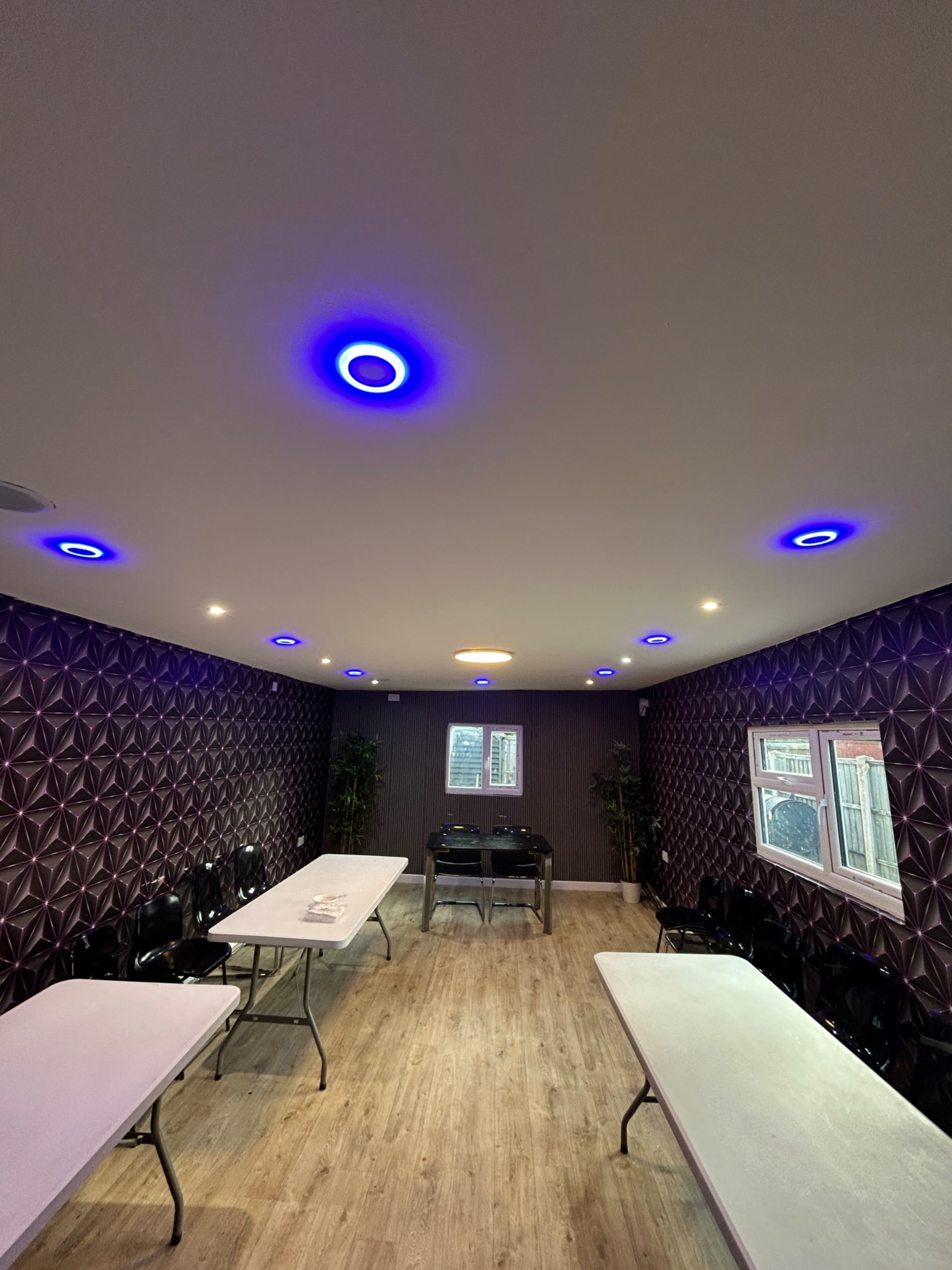 The image shows an indoor room with multiple rectangular tables arranged in the center and purple-lit circular ceiling fixtures above.