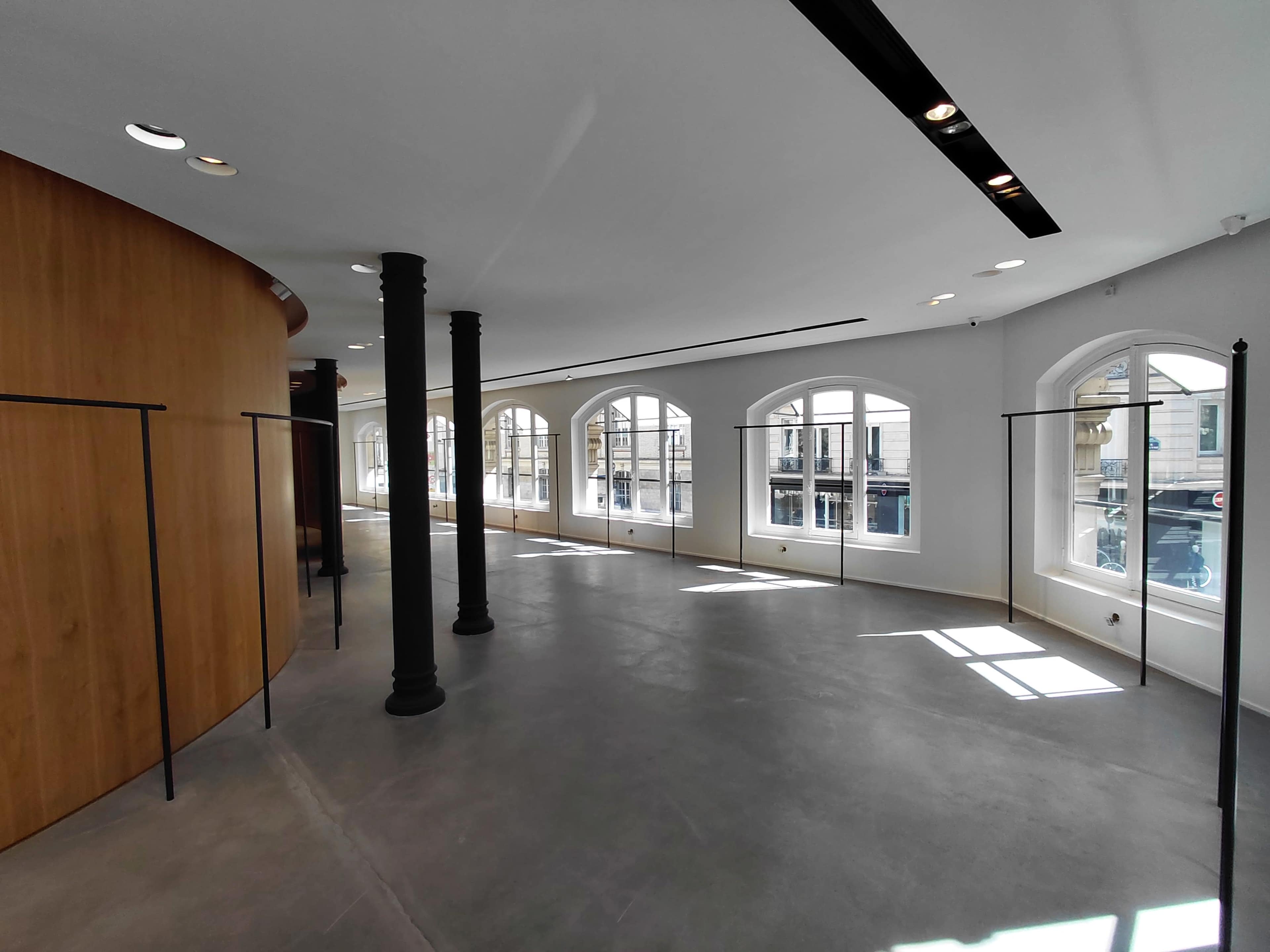 The image shows a spacious interior with large arched windows, a smooth concrete floor, and minimal clothing racks along the walls.