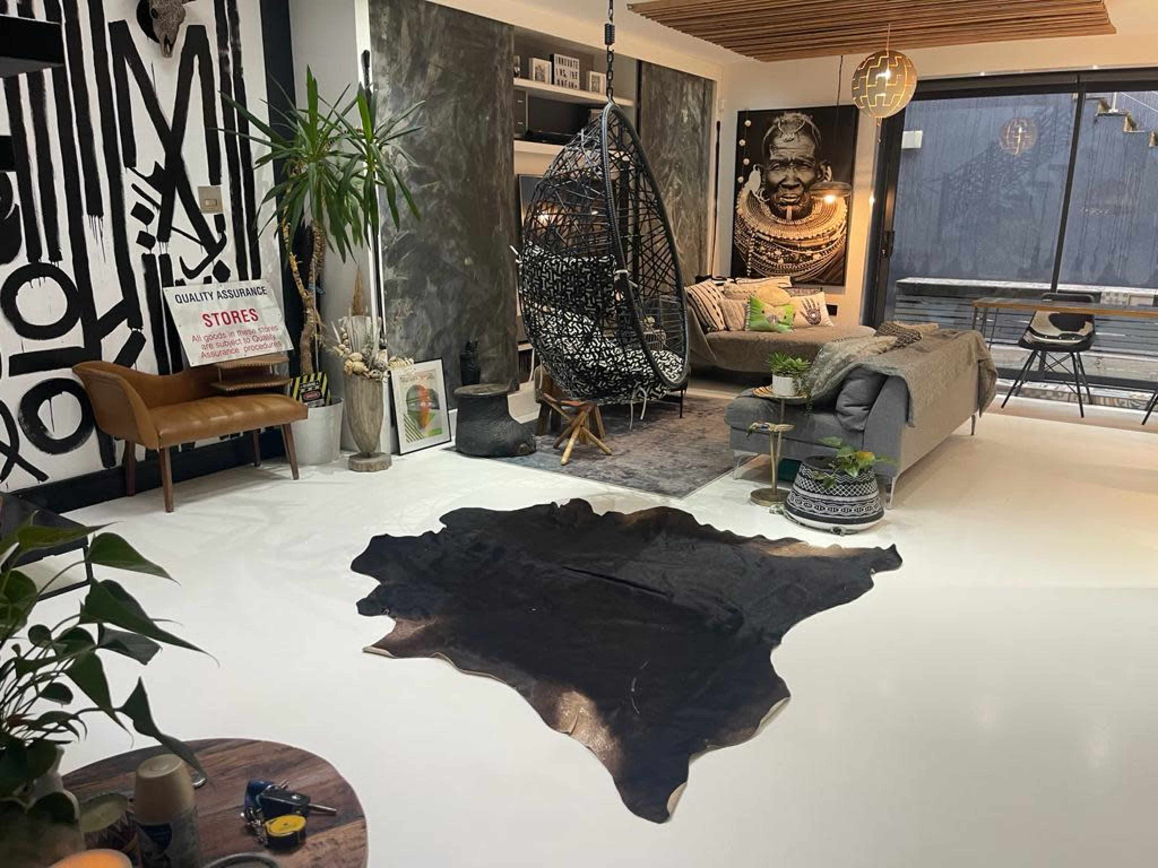 The room features a modern living space with a hanging wicker chair, a bed, a cowhide rug, and various indoor plants against a backdrop of decorative wall art.