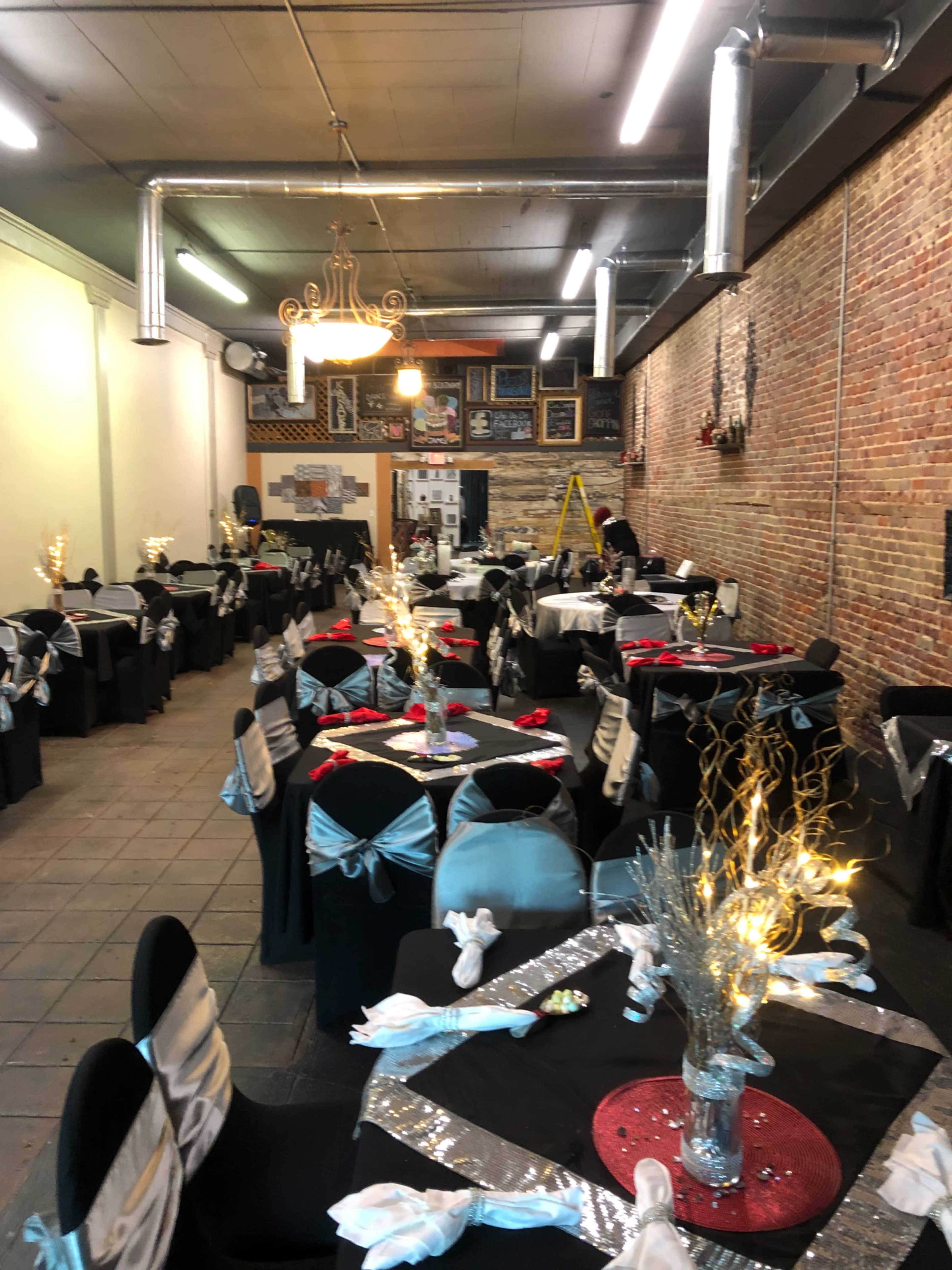 A spacious dining area set with numerous tables covered in black cloth, adorned with silver and blue decorations, and featuring centerpieces with lights.