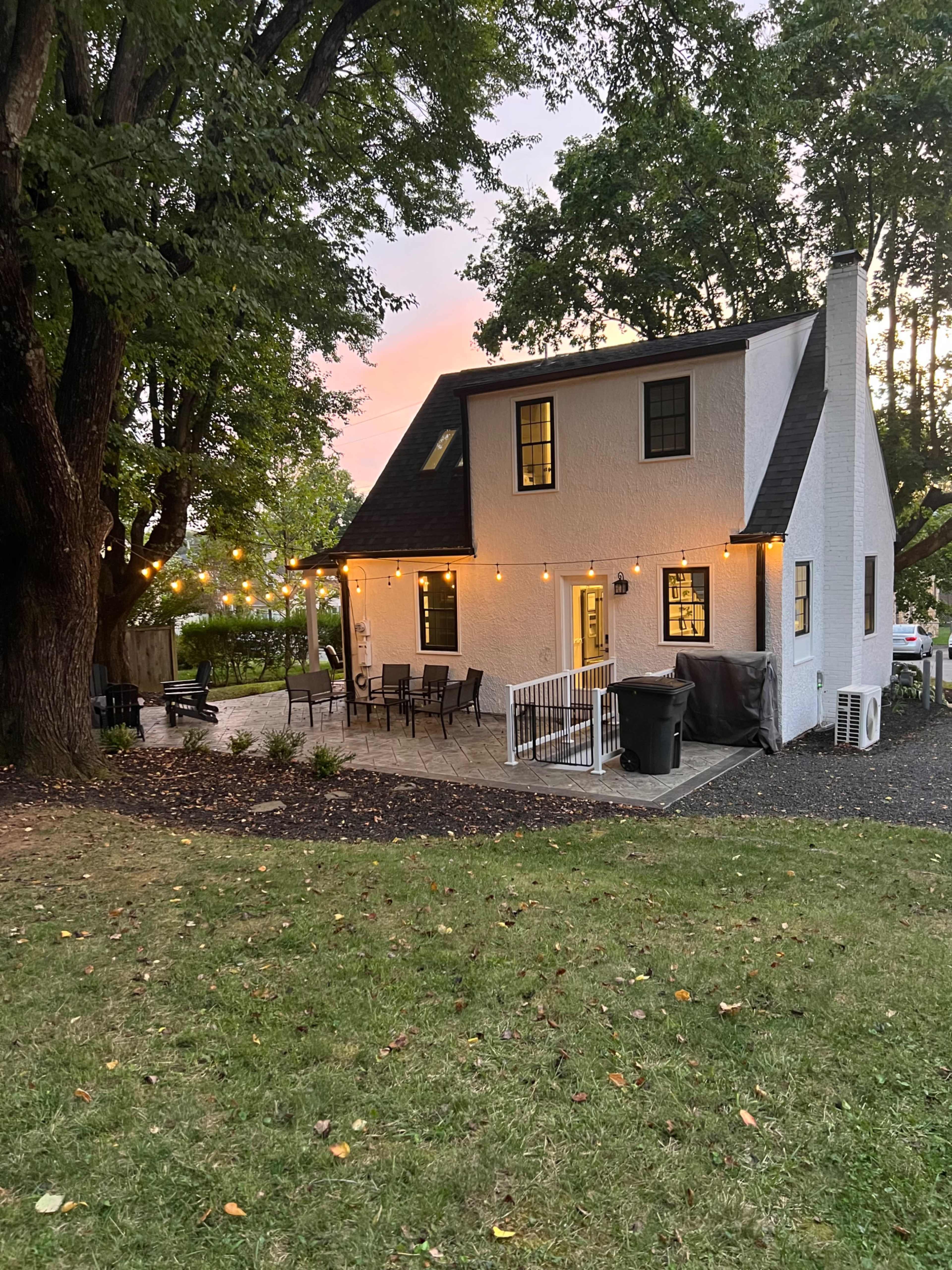 A white house with a sloping roof and outdoor seating area is illuminated by string lights, surrounded by a grassy yard and trees during sunset.