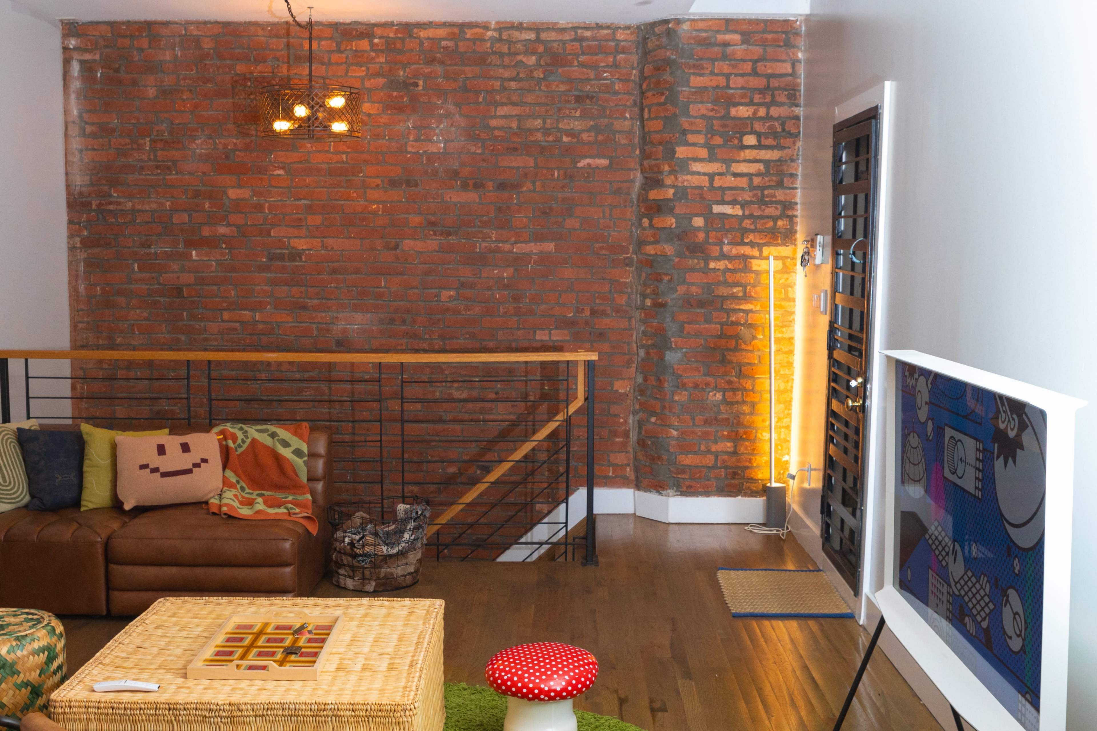A modern living room with exposed brick walls, a brown leather sofa, a wicker basket, and a colorful accent rug.