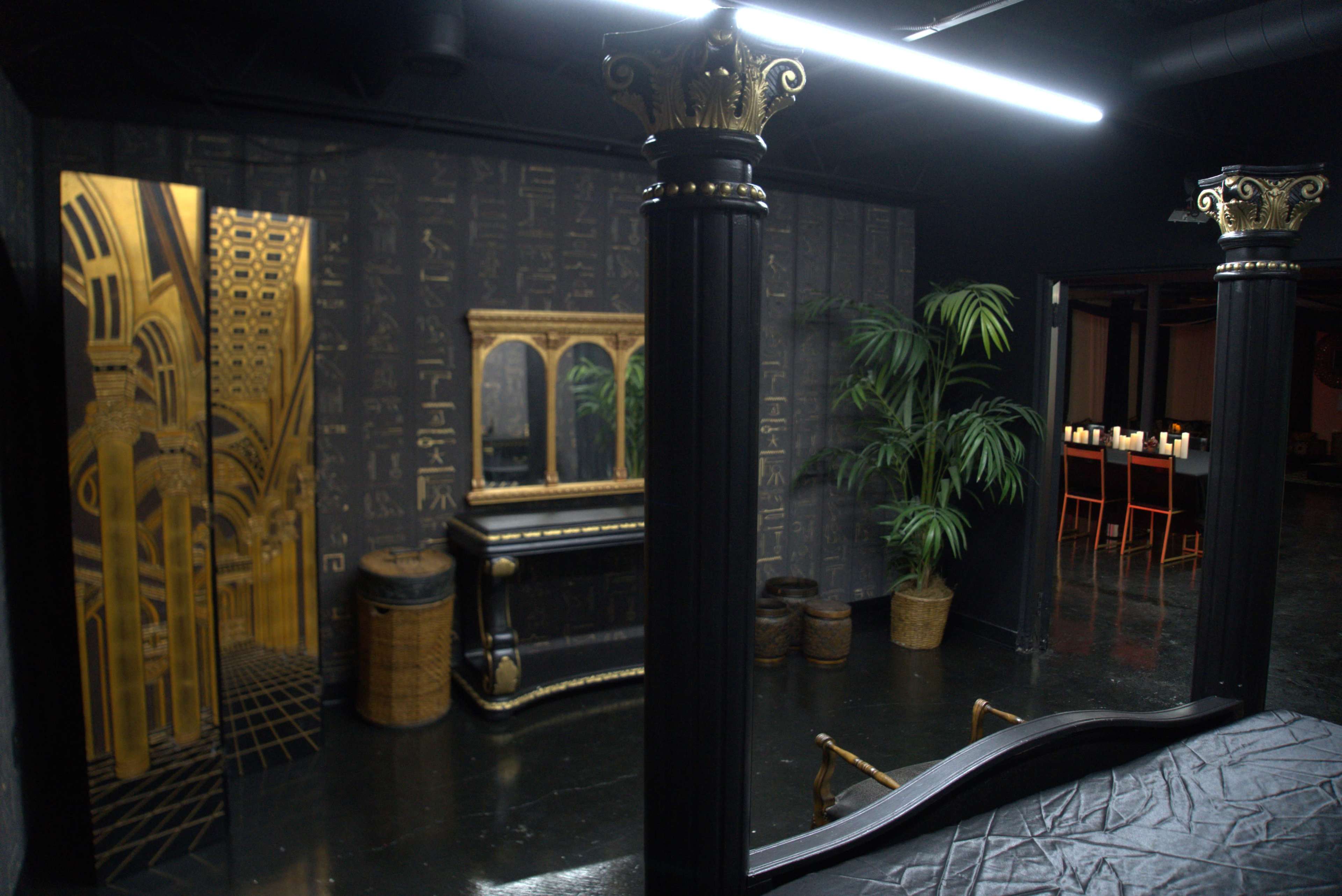 An elegantly decorated room featuring black pillars, a mirrored dresser, a gold accent wall with an architectural design, and potted plants.