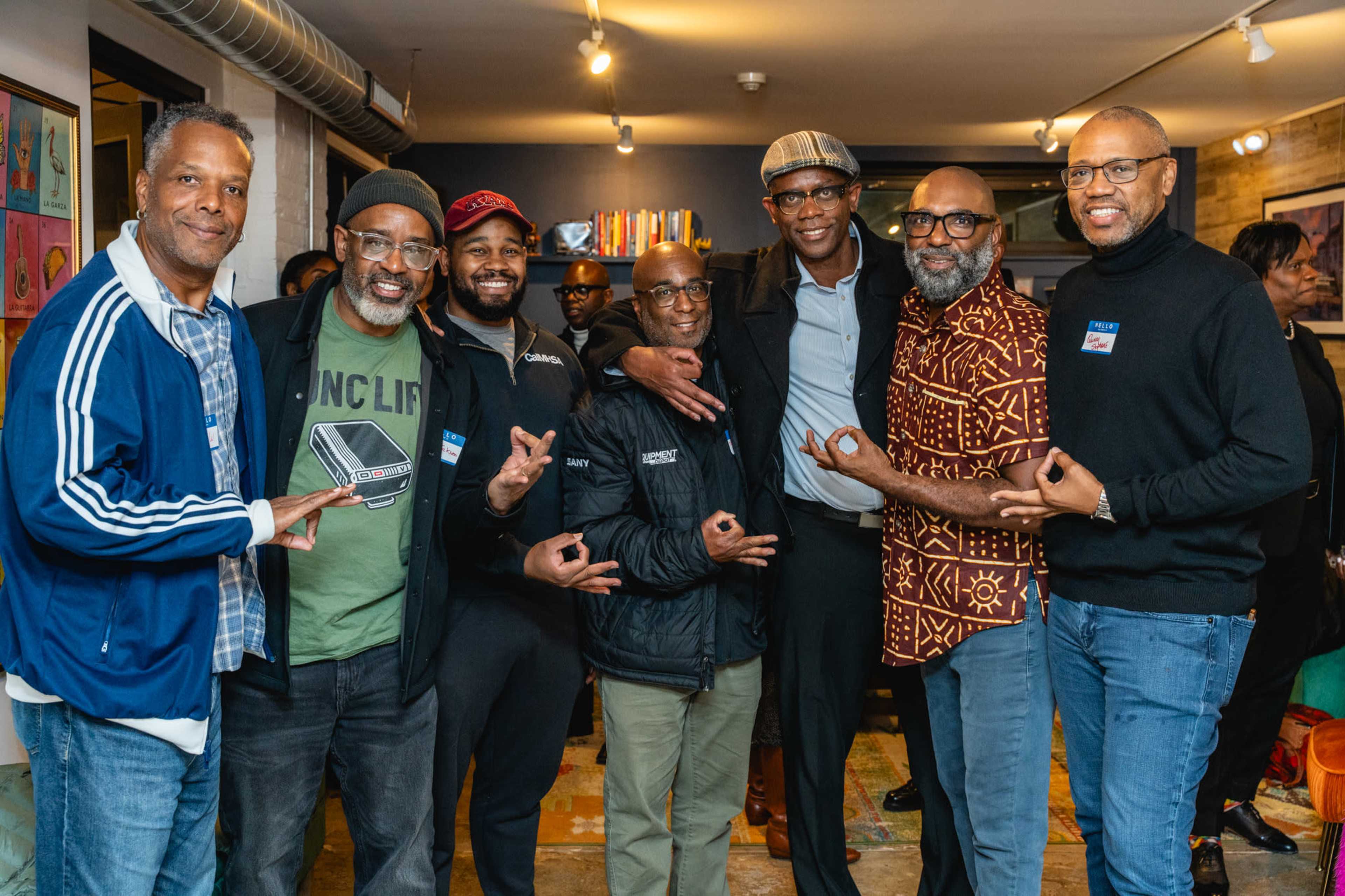 A group of seven men stands together in a casual indoor setting, posing for a photo with smiles and hand signals.