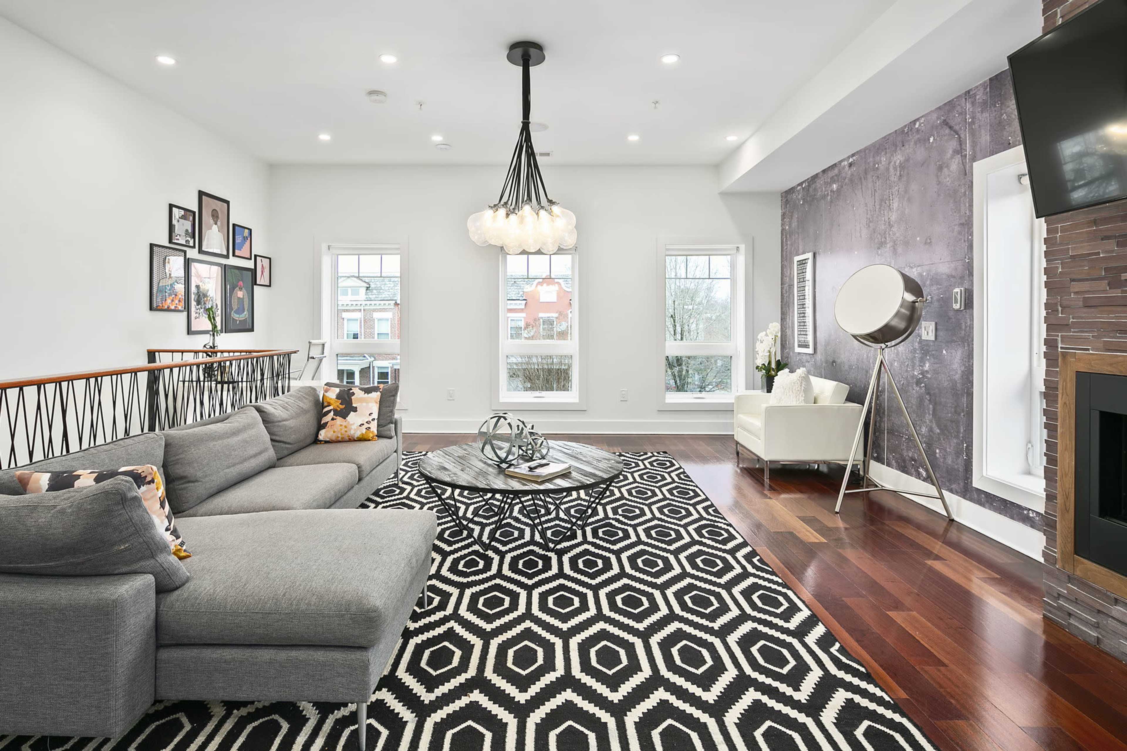 The living room features a gray sectional sofa, a geometric black and white patterned rug, and a modern chandelier hanging above a circular coffee table.