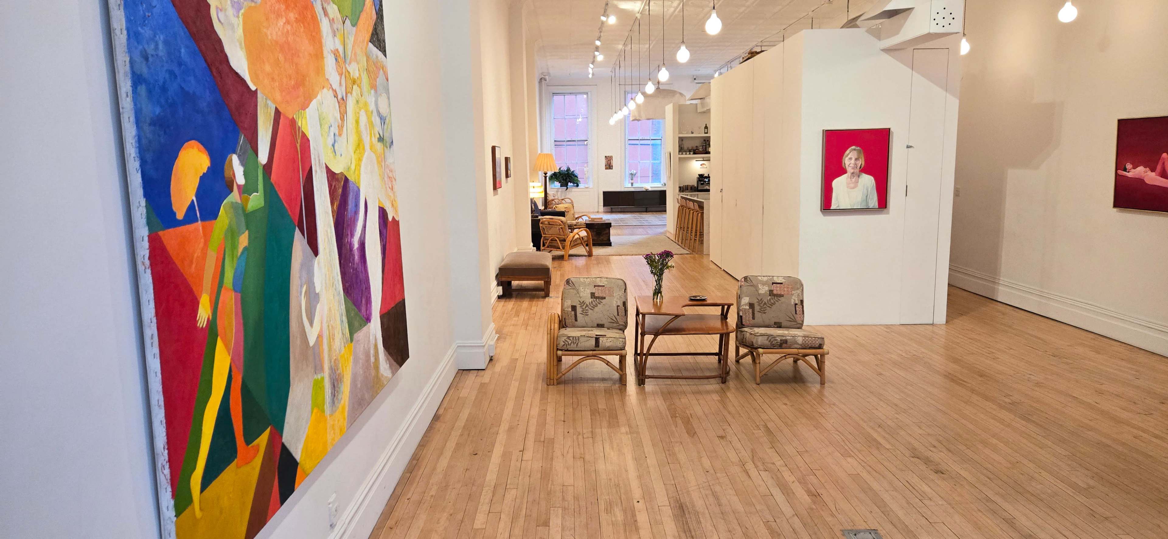 The image shows a spacious art gallery with colorful abstract artwork on the left wall, light-colored wooden flooring, and a seating area with two armchairs in the foreground.