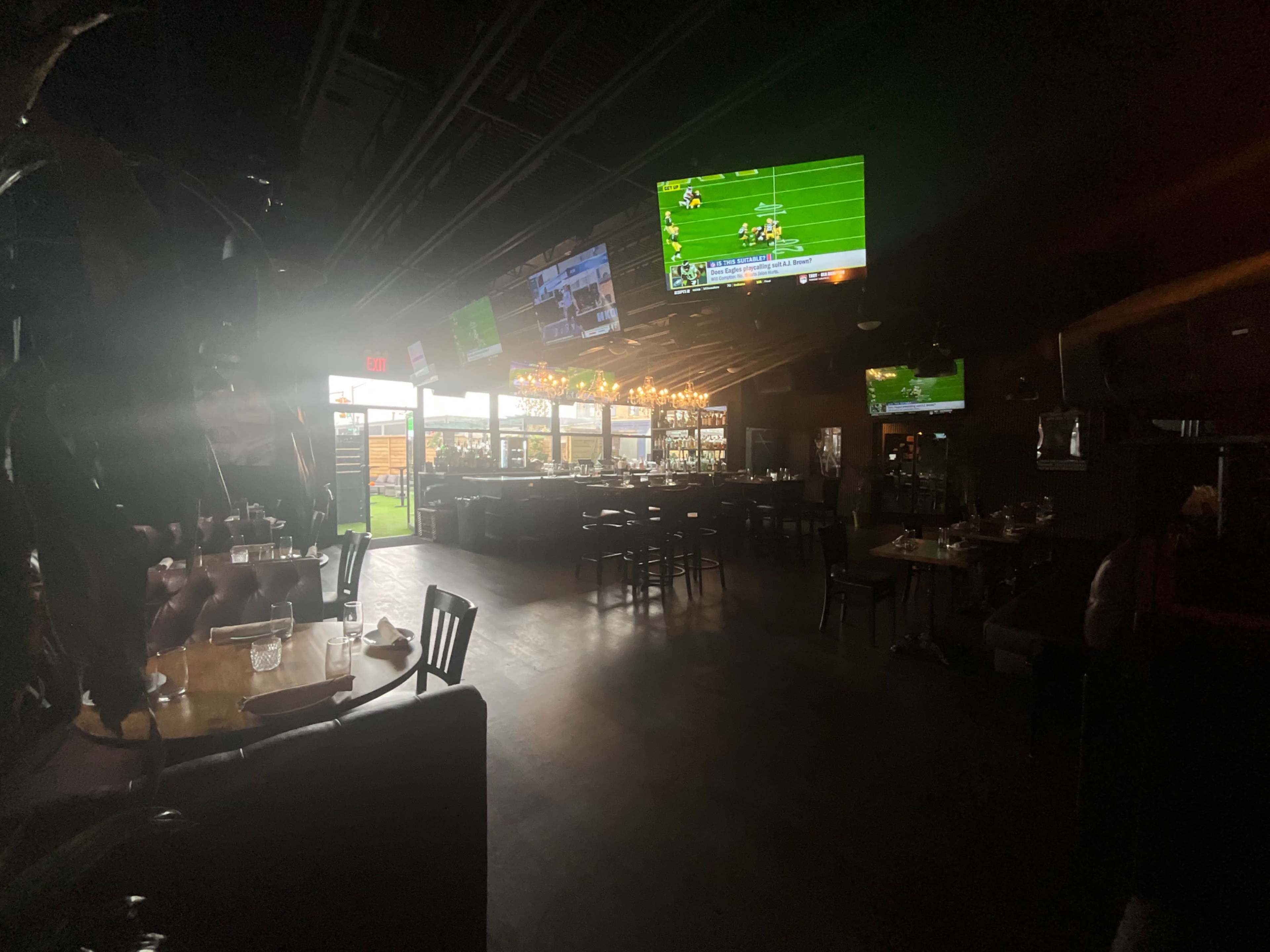 The image shows a dimly lit restaurant interior with several televisions displaying sports and an open entrance leading to an outdoor area.