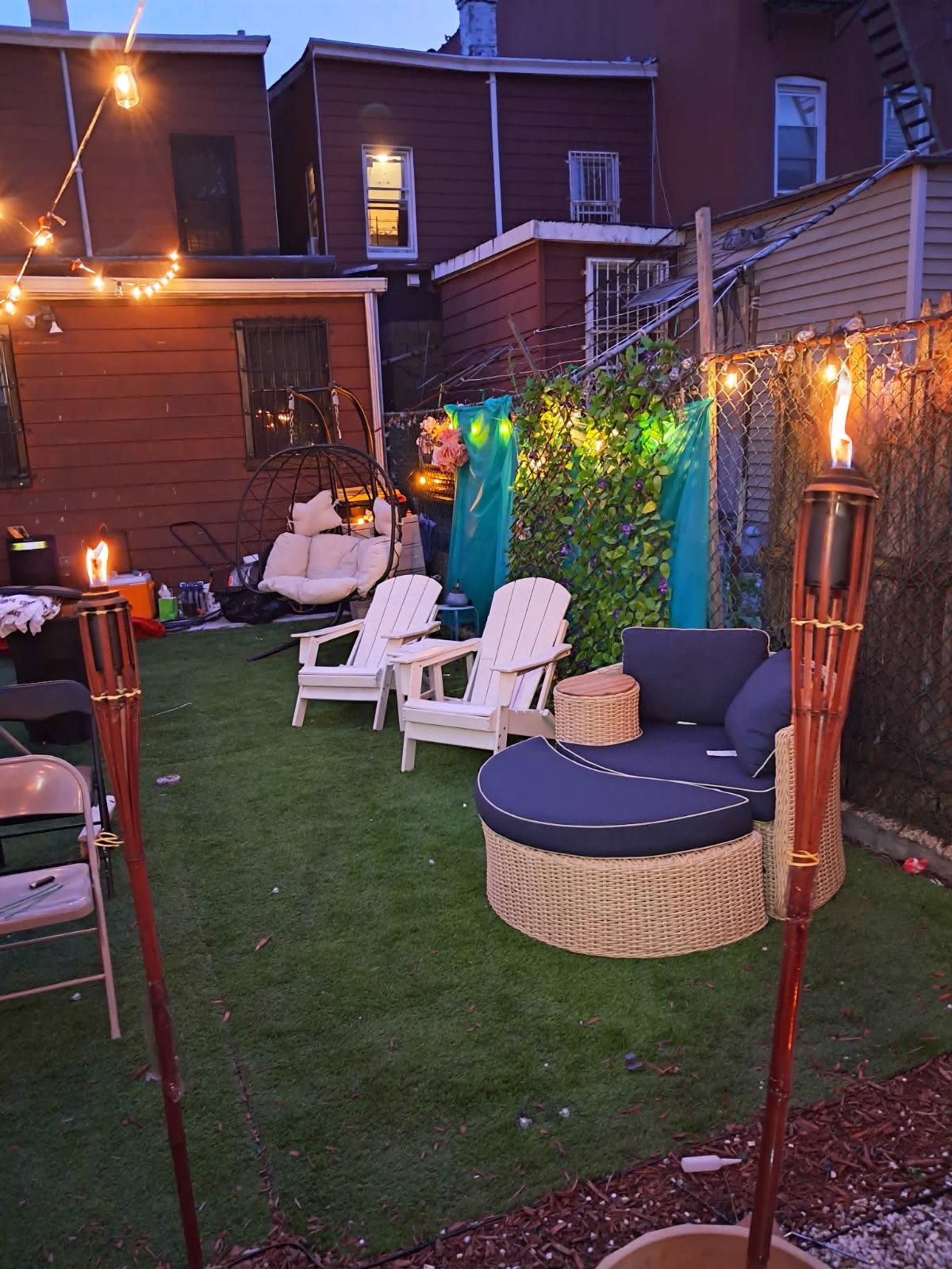 The image shows a small outdoor space with two white Adirondack chairs and a round wicker lounge chair, surrounded by string lights and tiki torches.