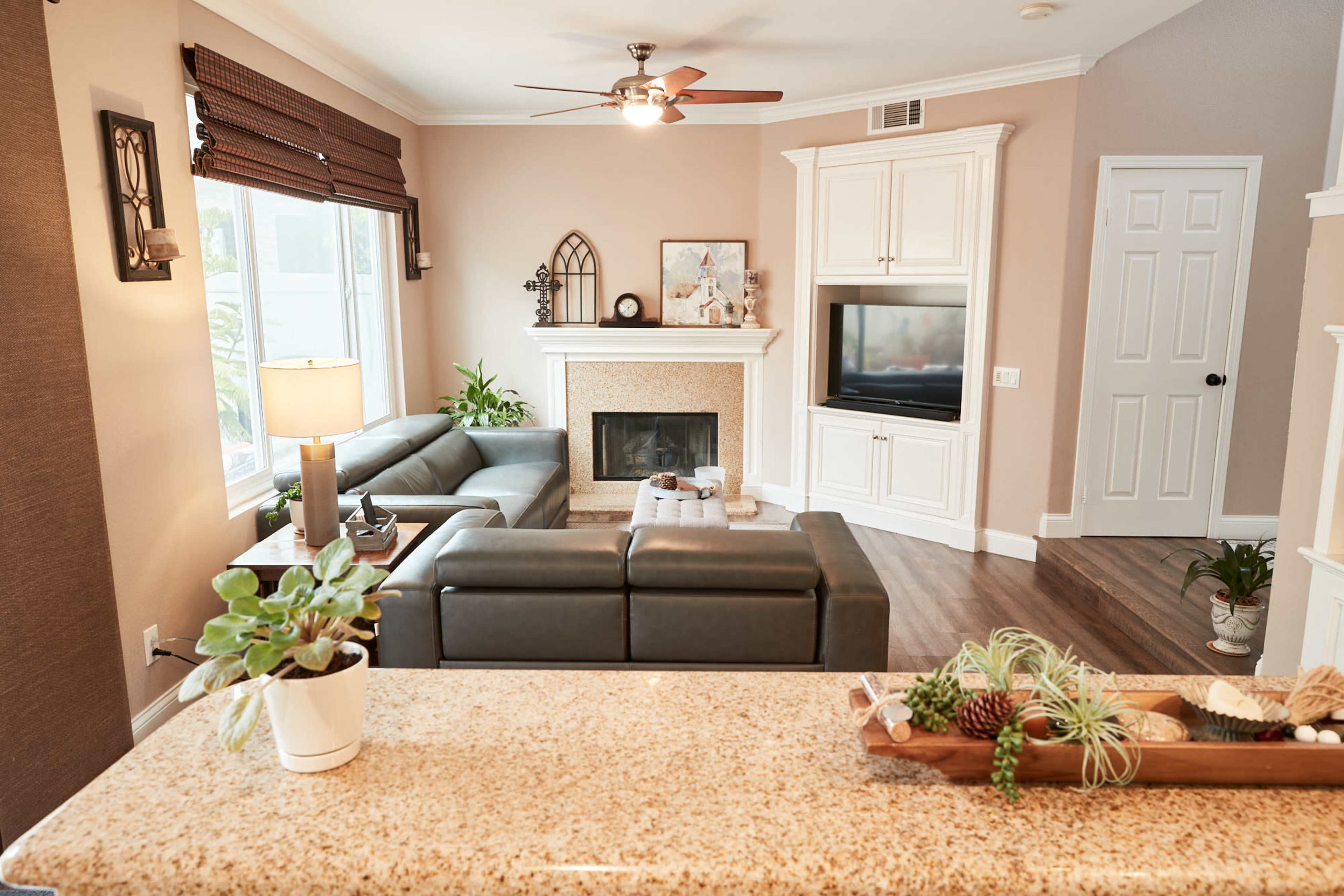 A living room with a gray sectional sofa, a fireplace, a television in a white cabinet, and a light-colored countertop in the foreground.