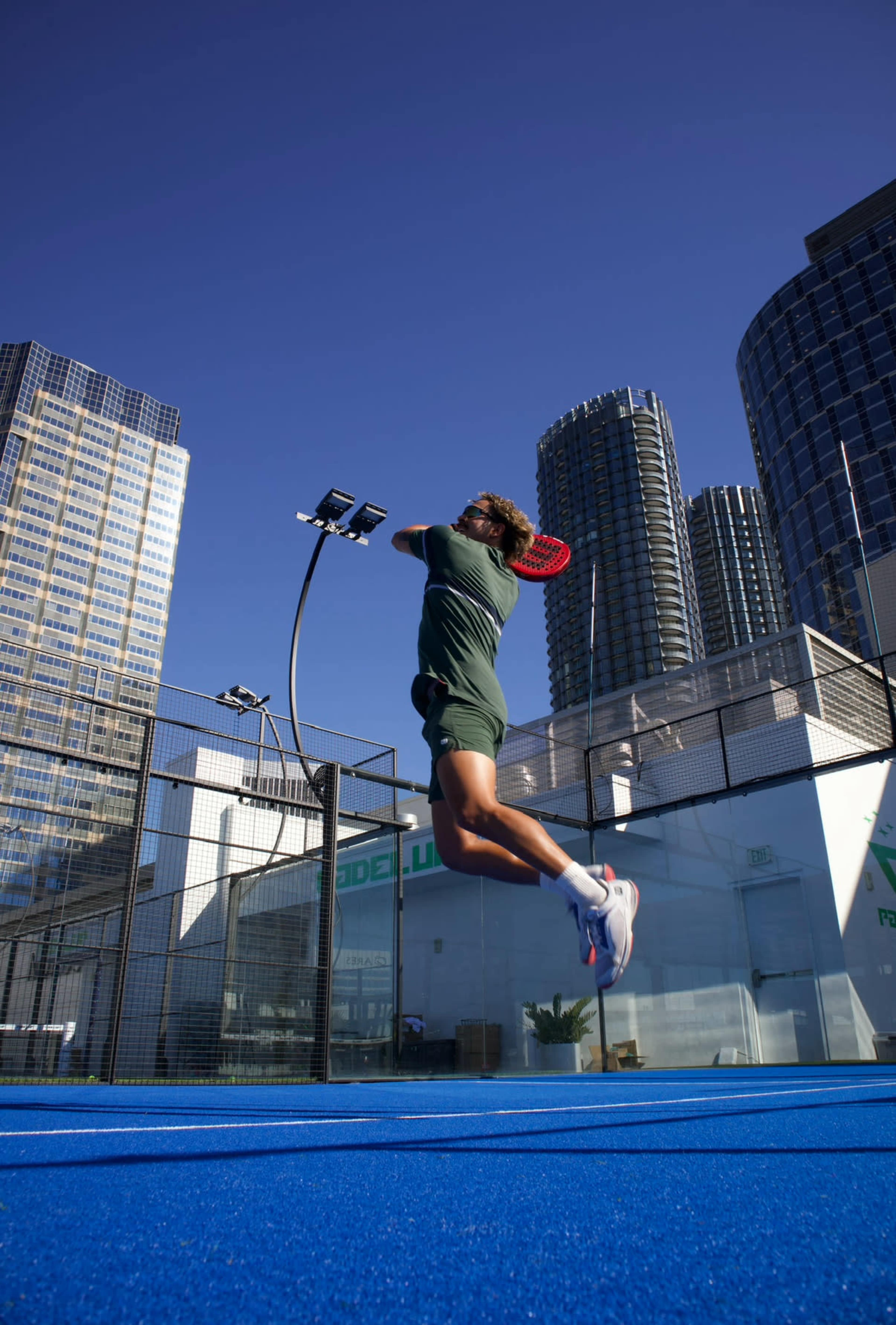 A player leaps in mid-air to hit a tennis ball on a blue court surrounded by tall buildings.