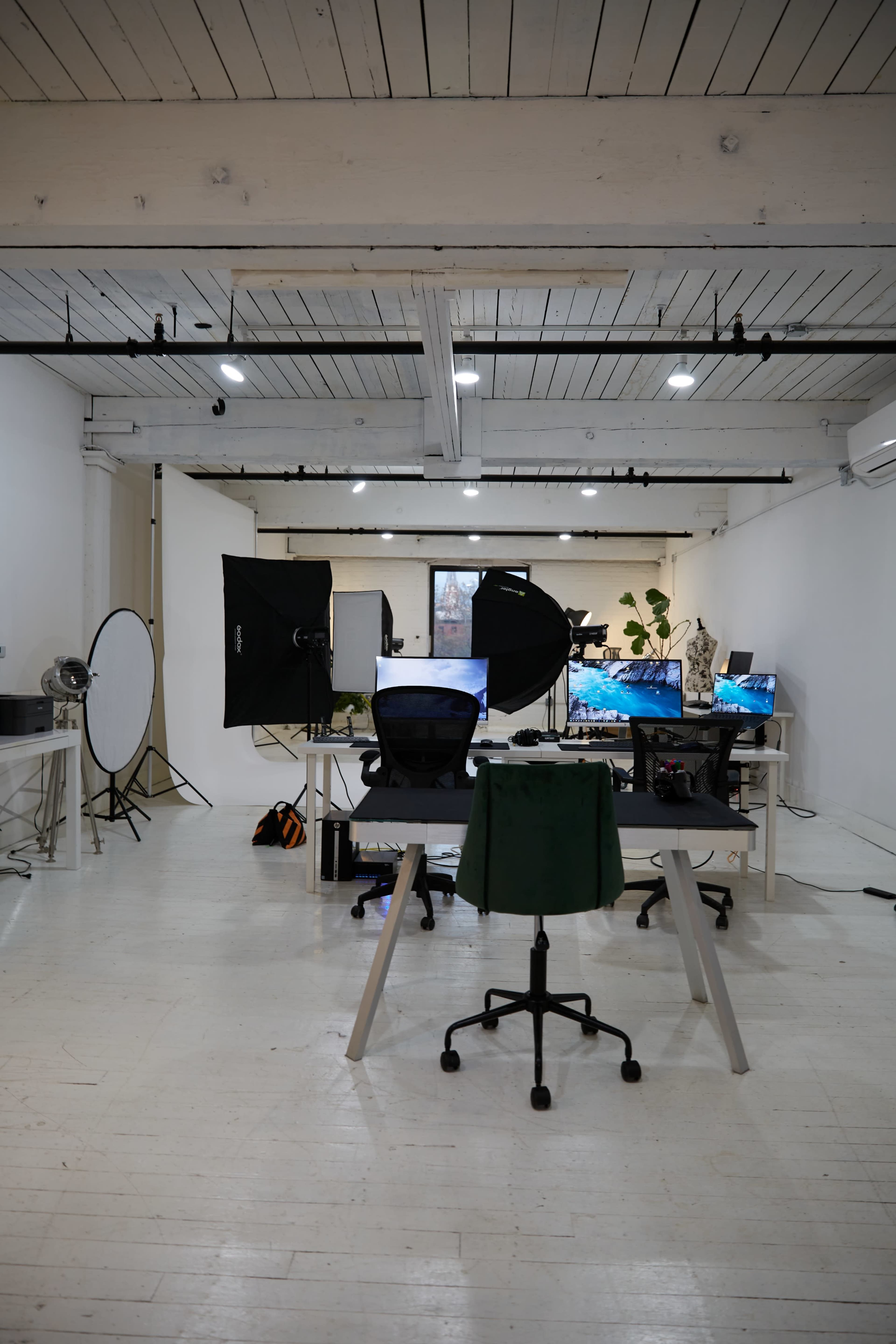 A bright, minimalistic studio space features multiple computer monitors, a green table, and photography equipment set up against white walls.
