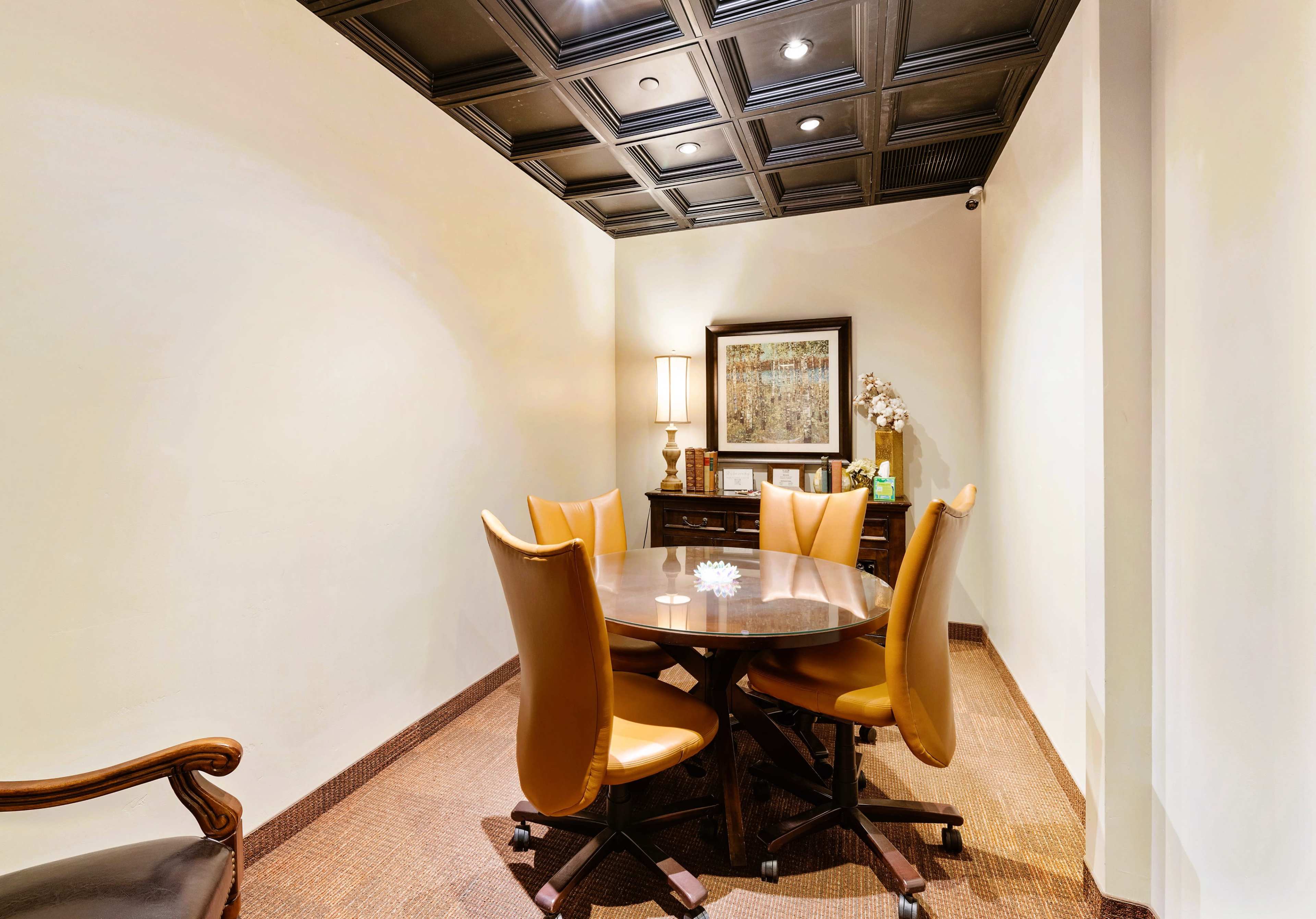 A small conference room features a round table surrounded by four brown leather chairs, with a lamp and artwork on the wall.