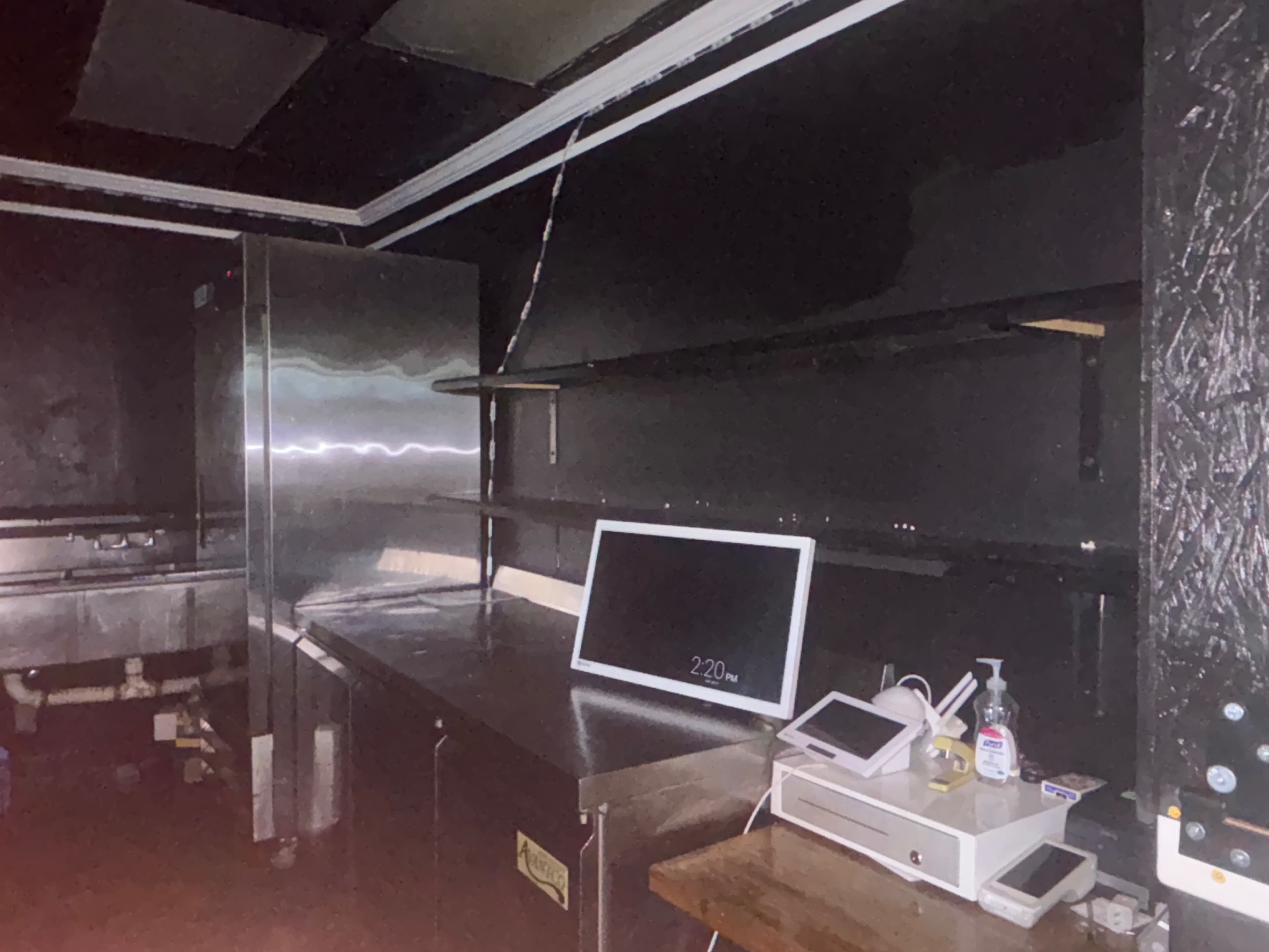 The image shows a dimly lit commercial kitchen with stainless steel appliances, empty shelves, and a computer monitor on a wooden table.
