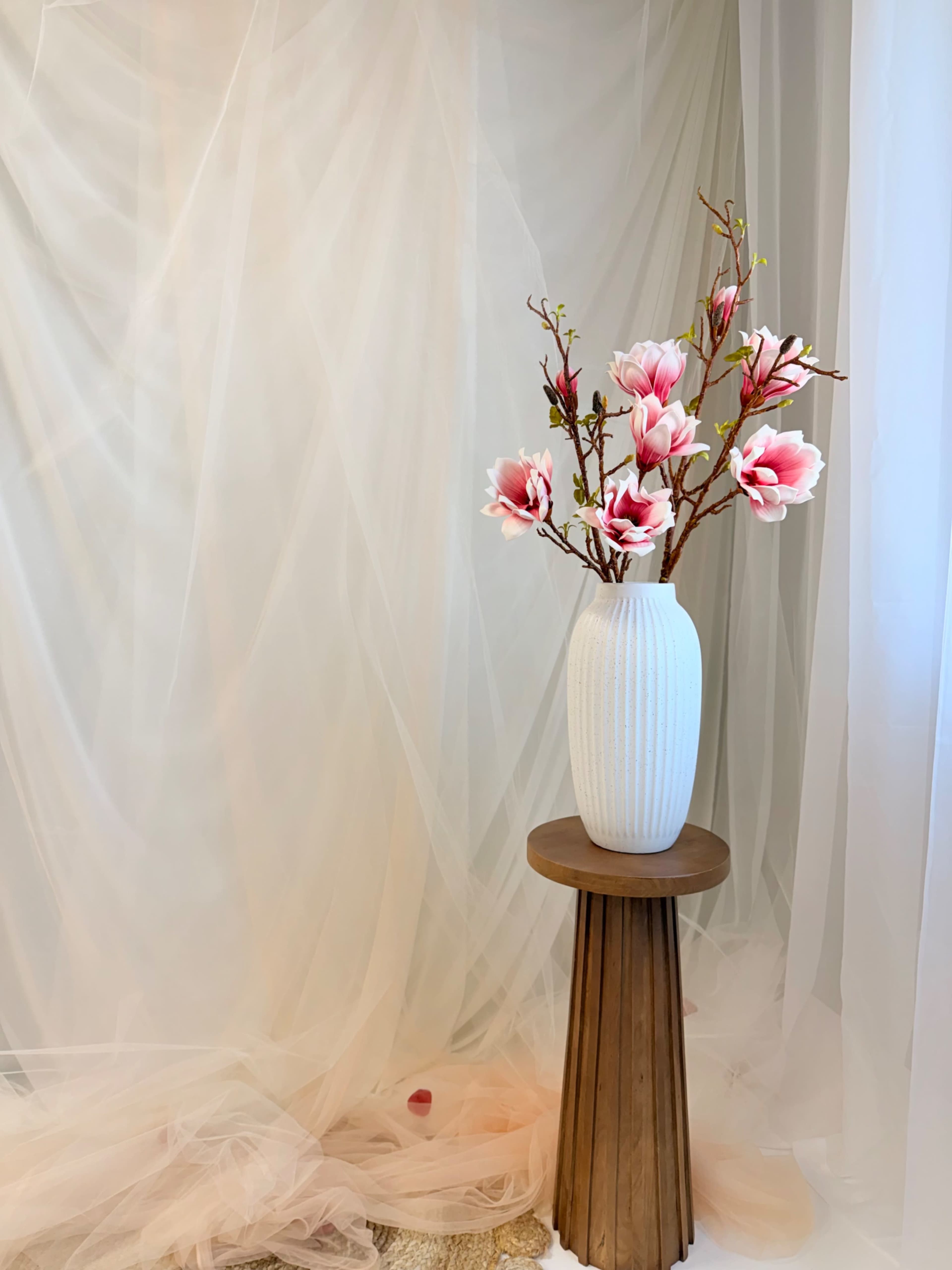 A white vase filled with pink flowers stands on a wooden pedestal in front of a draped, sheer background.