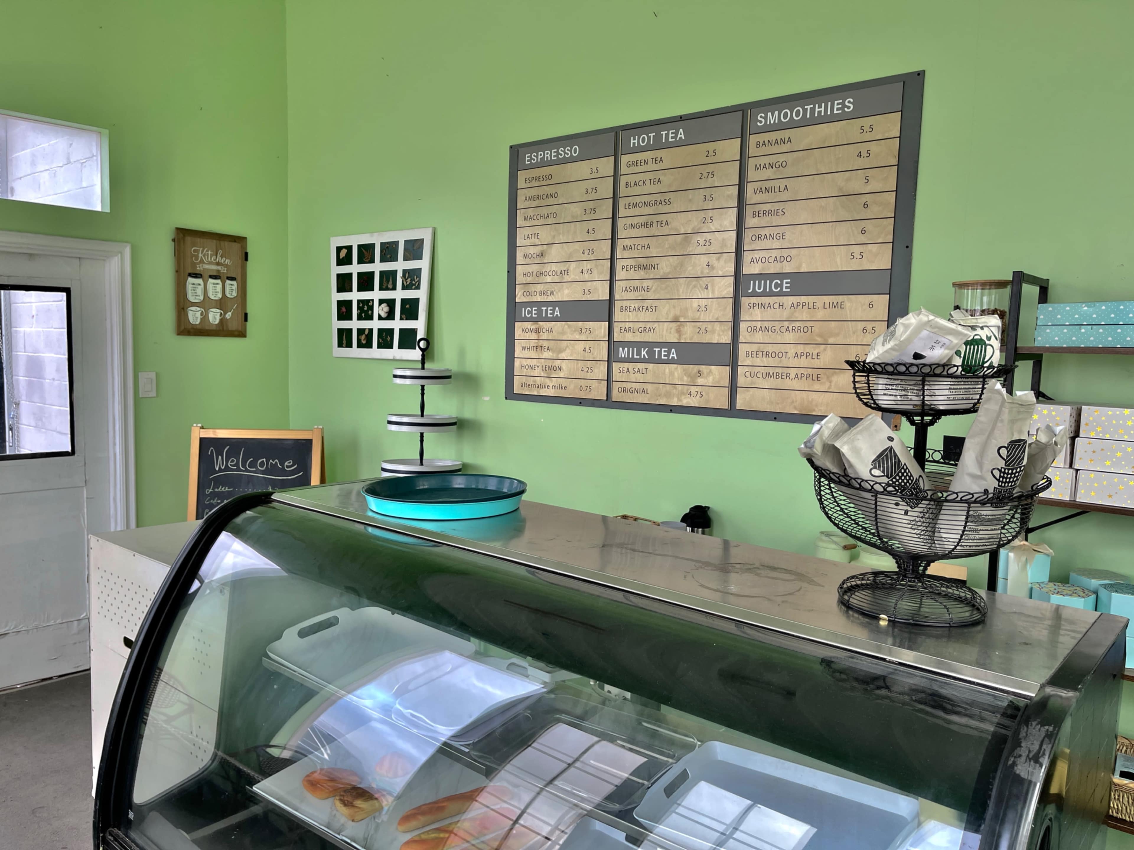 The image shows a cafe interior with a green wall, a refrigerated display case, and a menu board listing various beverages and snacks.