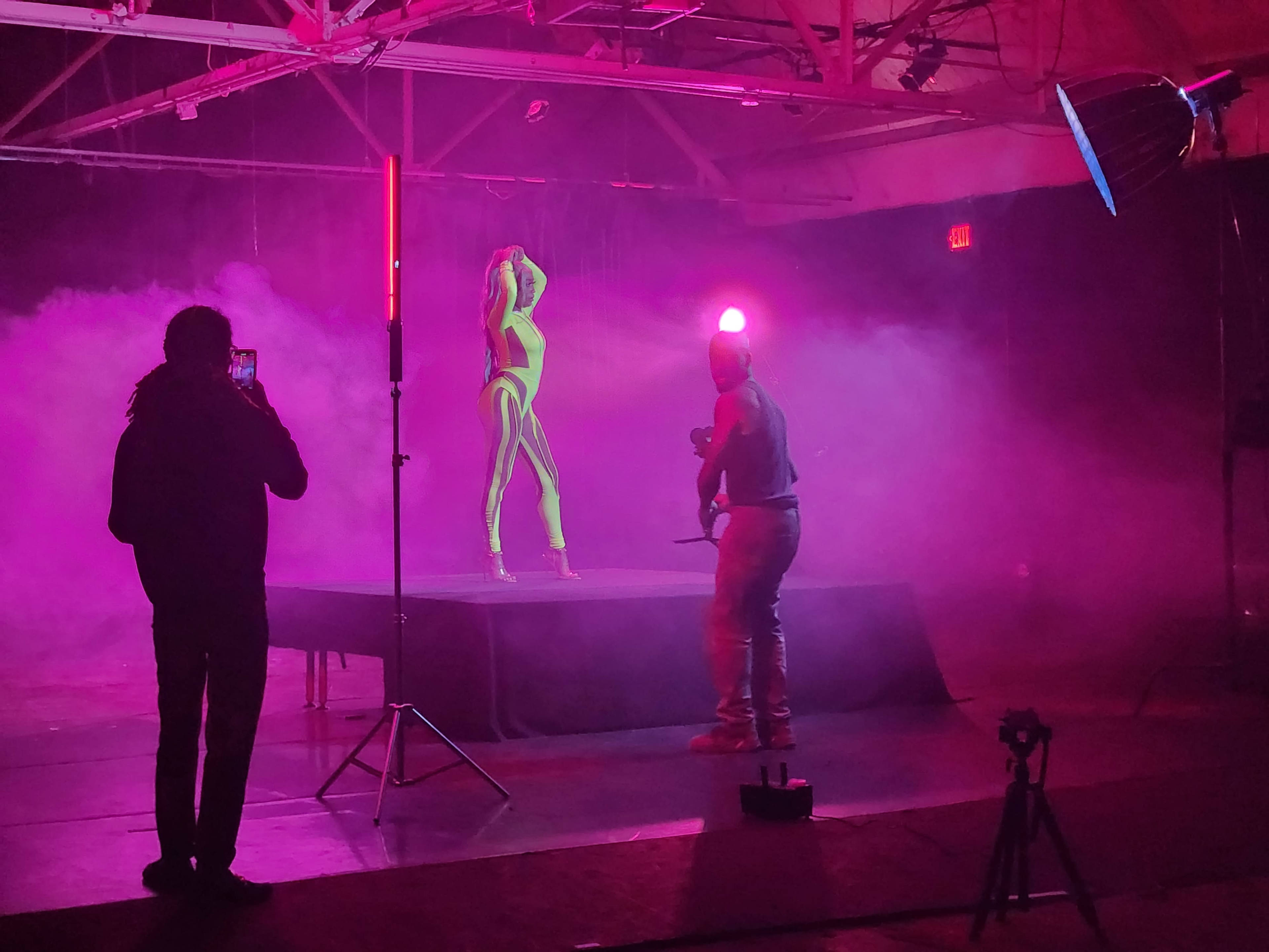 A model poses on a platform while a photographer captures images in a studio filled with colored smoke and dramatic lighting.