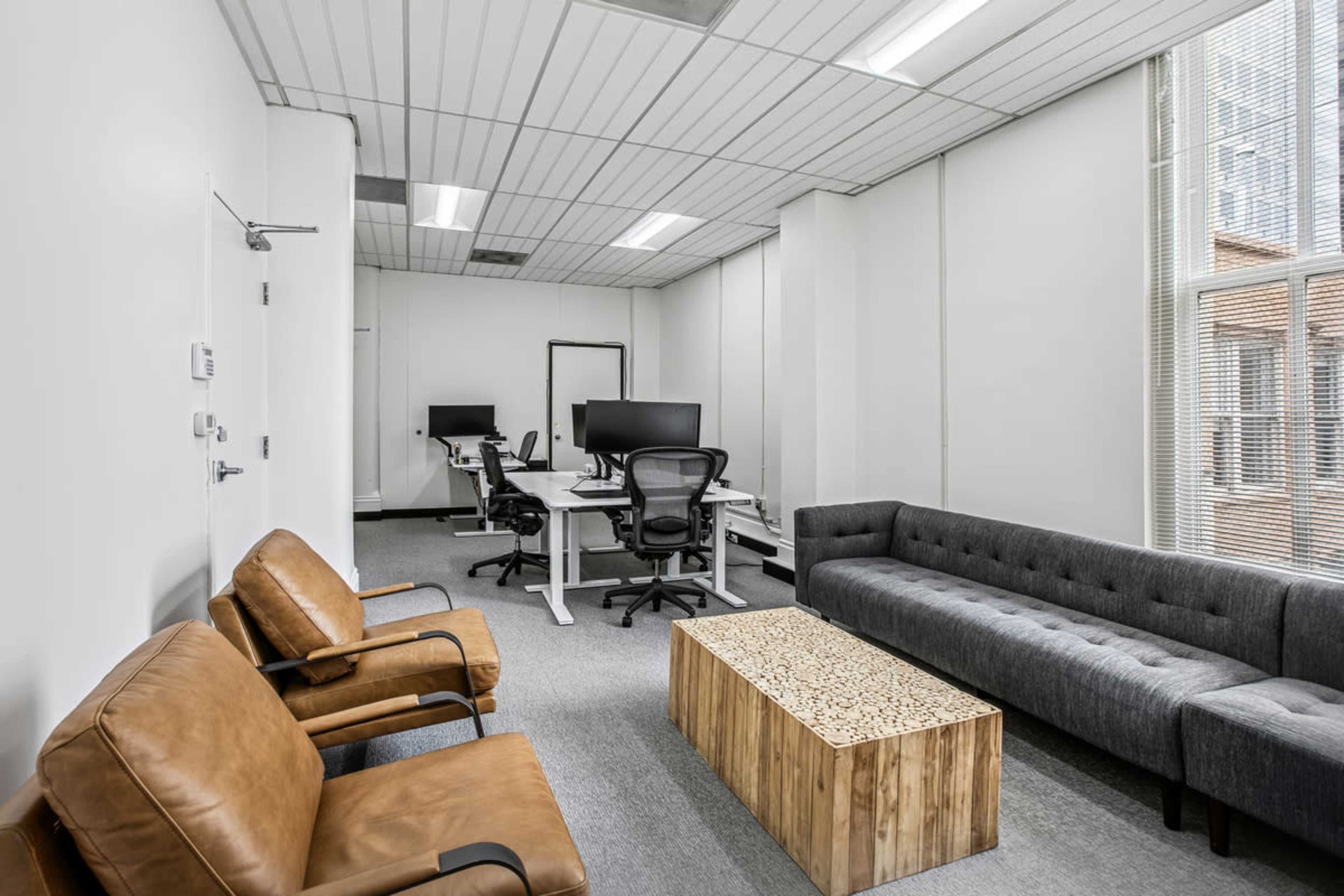 The image shows a modern office space featuring two leather chairs, a sectional sofa, a wooden coffee table, and several workstations with dual monitors.