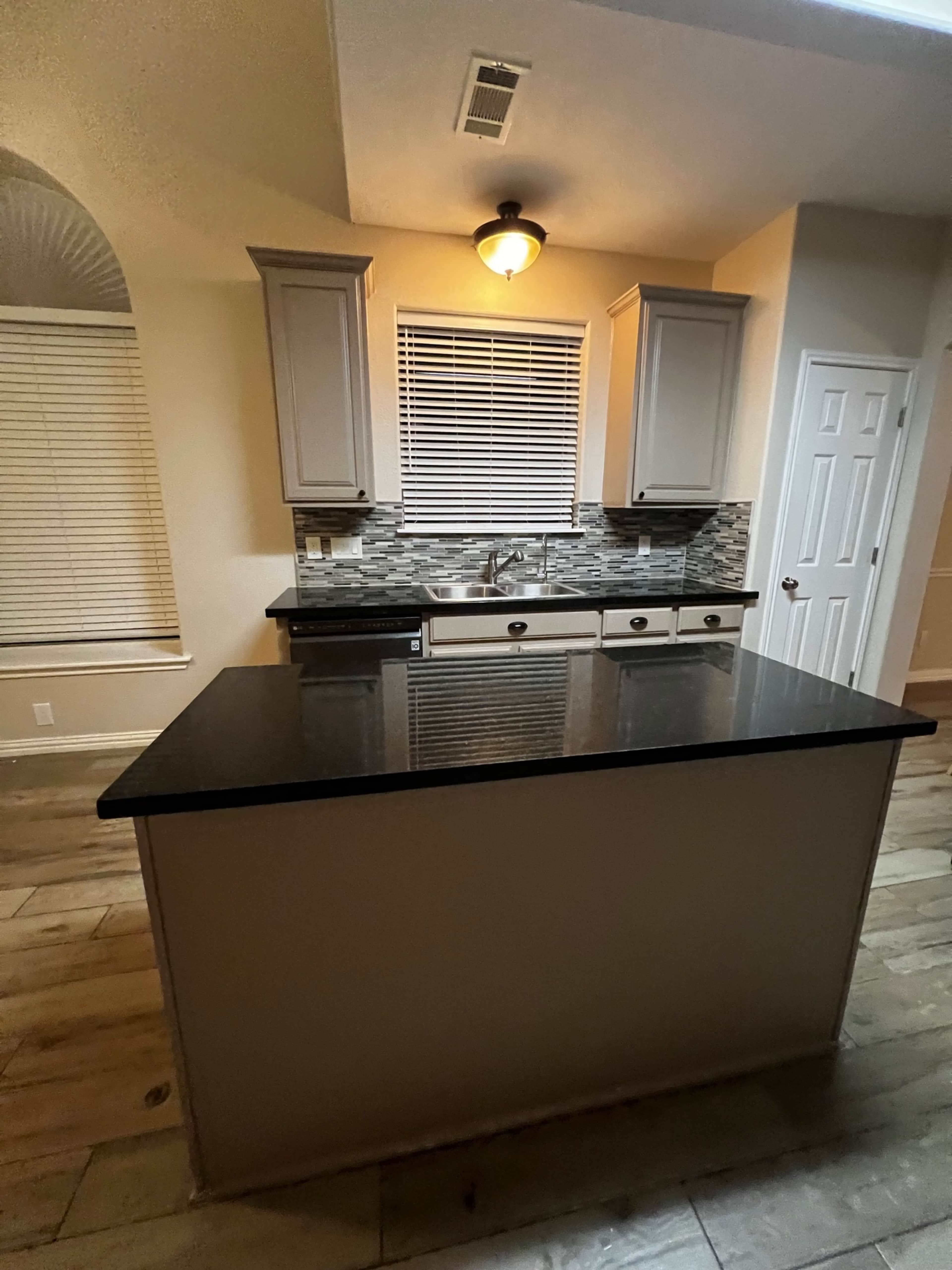 A kitchen features light gray cabinets, a black countertop, and a tiled backsplash with a window and blinds above the sink.