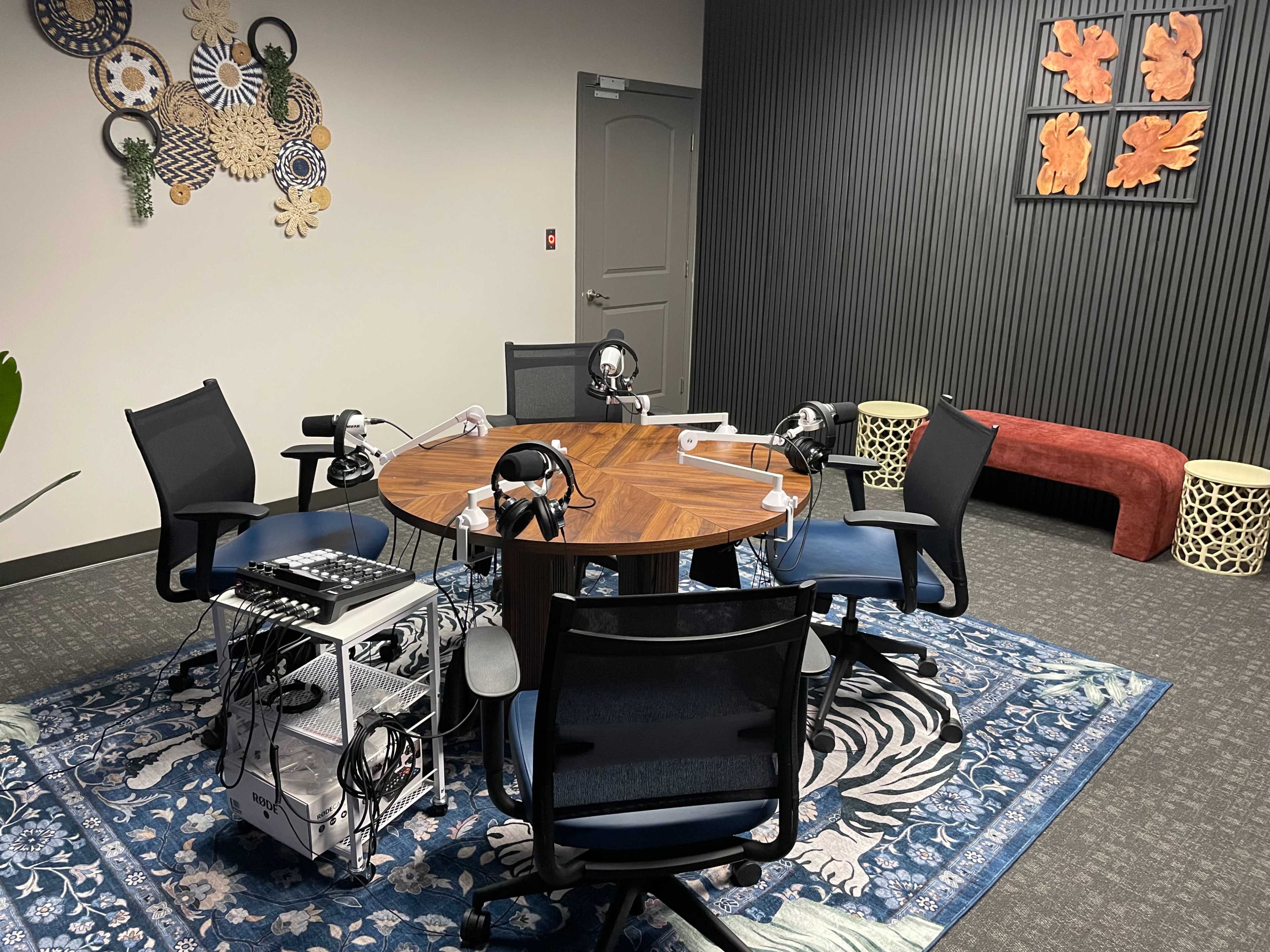The image shows a recording studio setup featuring a round wooden table surrounded by several black and blue chairs, with microphones and audio equipment arranged on the table.