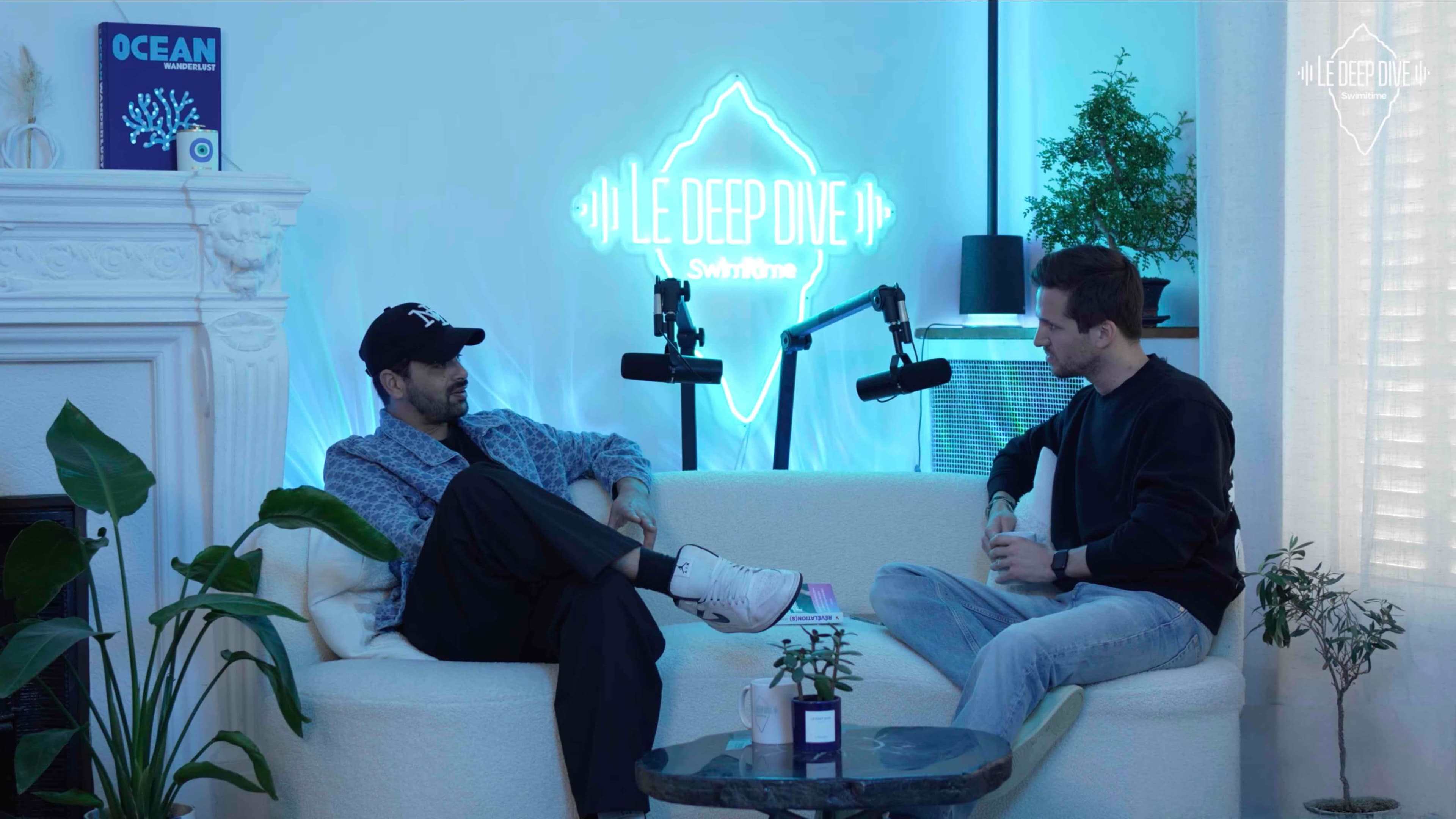 Two men sit on a white couch in a brightly lit room, with microphones positioned in front of them and a neon sign in the background.