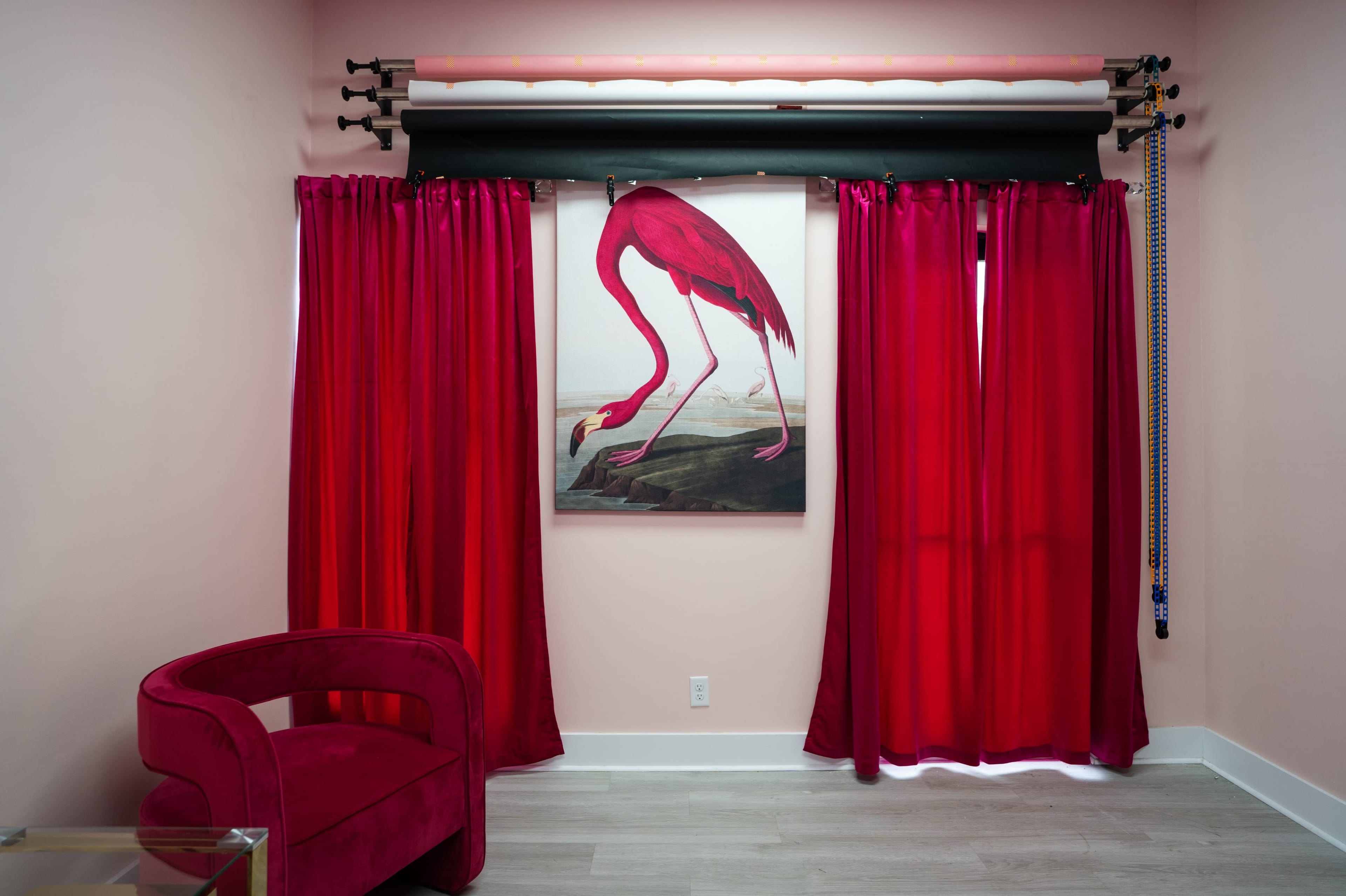 A room features a large painting of a flamingo on the wall, flanked by red curtains, with a pink chair in the corner.