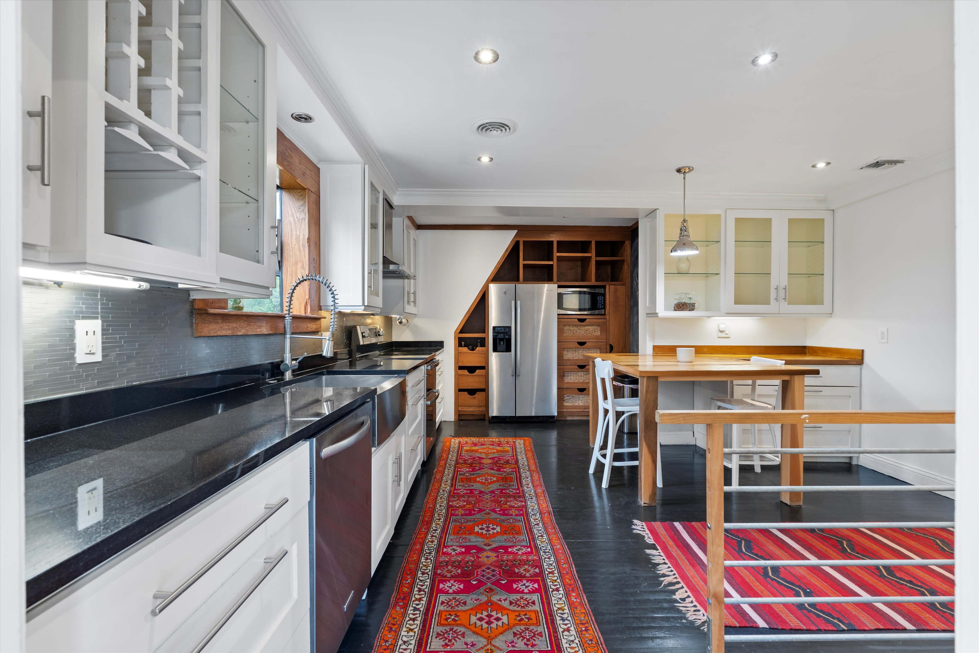 The kitchen features modern appliances, a central wooden table, and an area rug, with sleek cabinetry and a mix of dark countertops and light cabinetry.