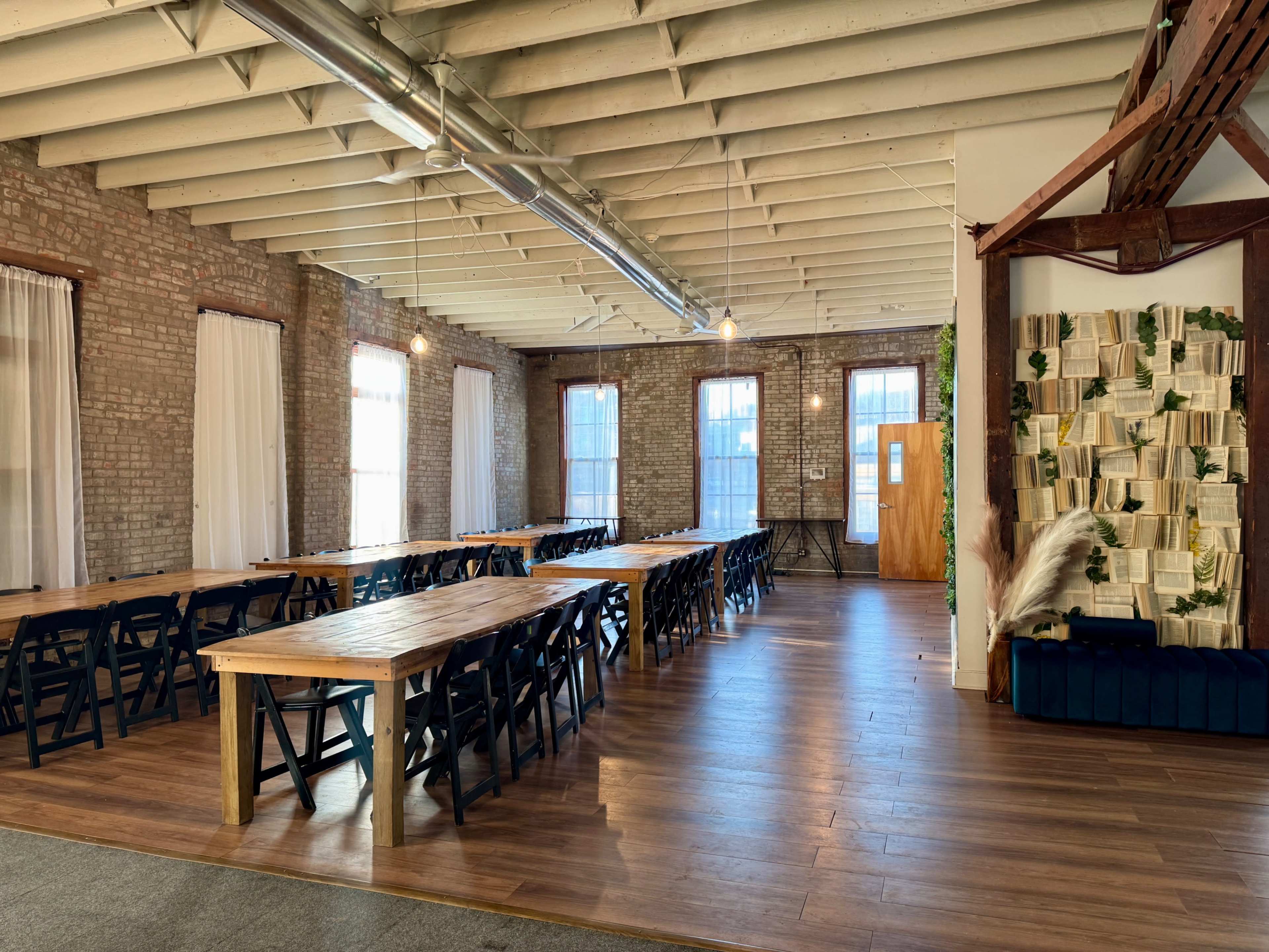 The image shows a spacious interior room with wooden tables arranged in a large U-shape, exposed brick walls, and several windows letting in natural light.