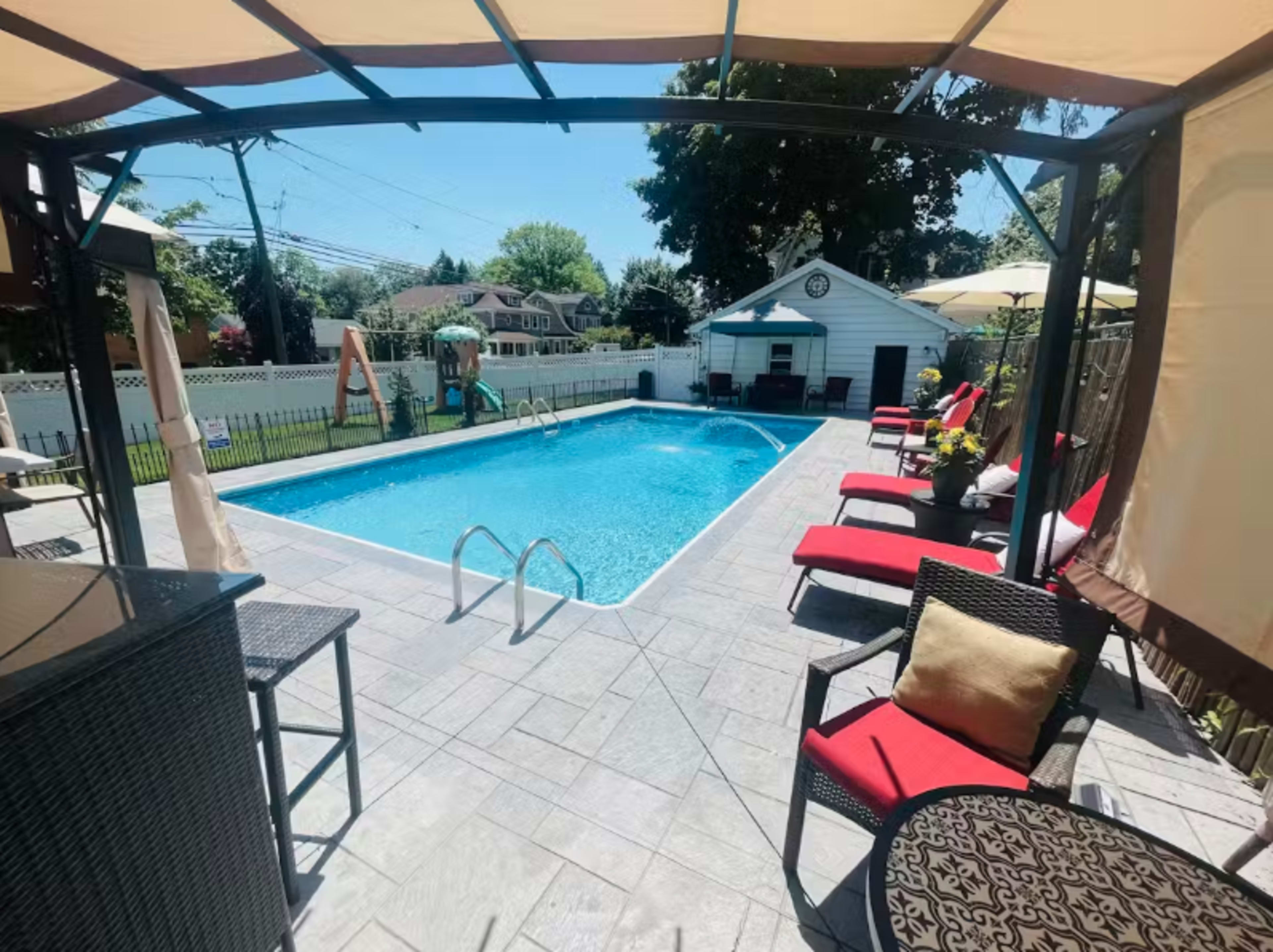 A swimming pool is surrounded by patio furniture, including red lounge chairs, and a gazebo in a residential backyard.