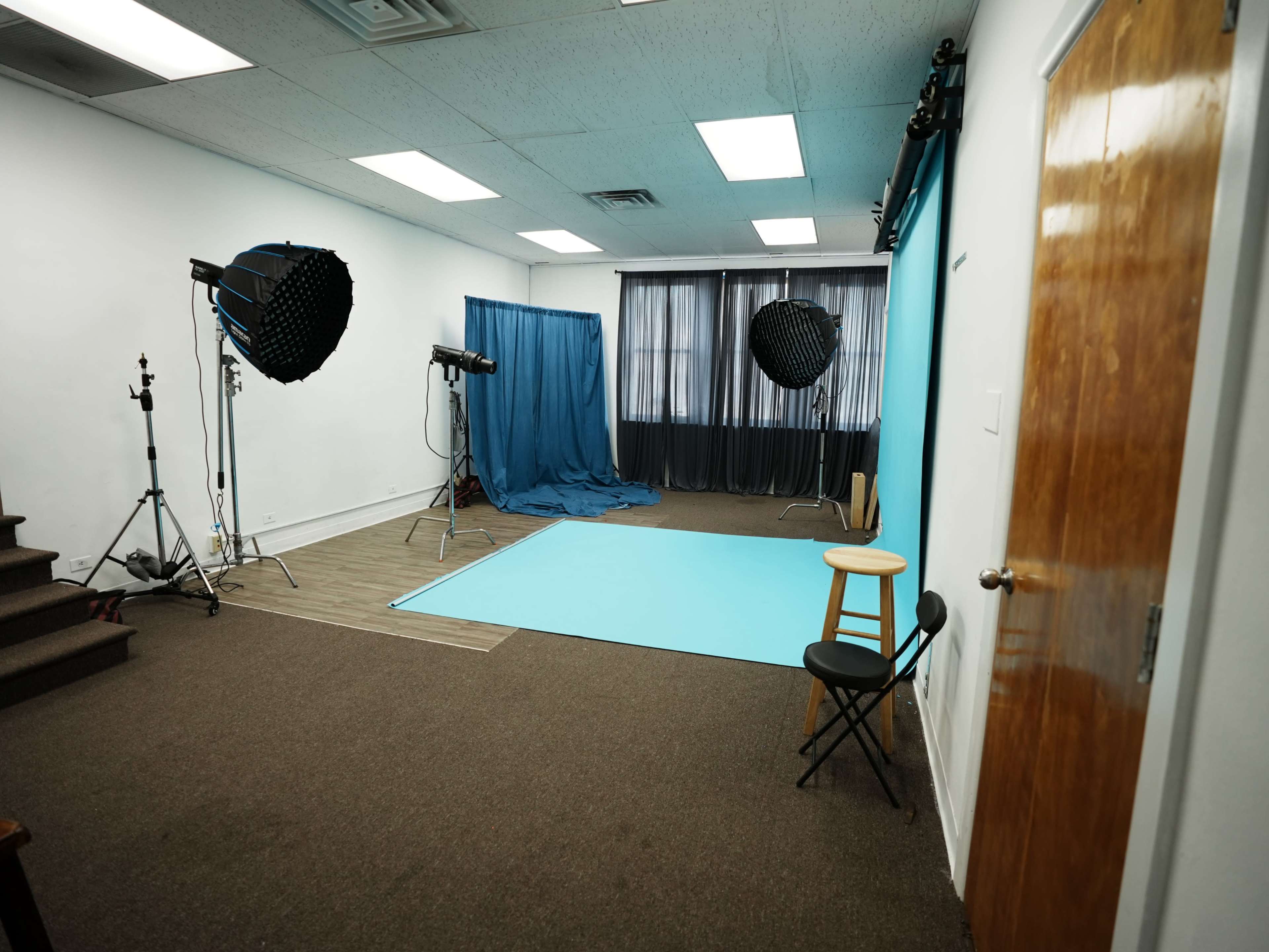 The image shows a photography studio featuring a blue backdrop, two light setups, a blue curtain, and a stool positioned in the corner.