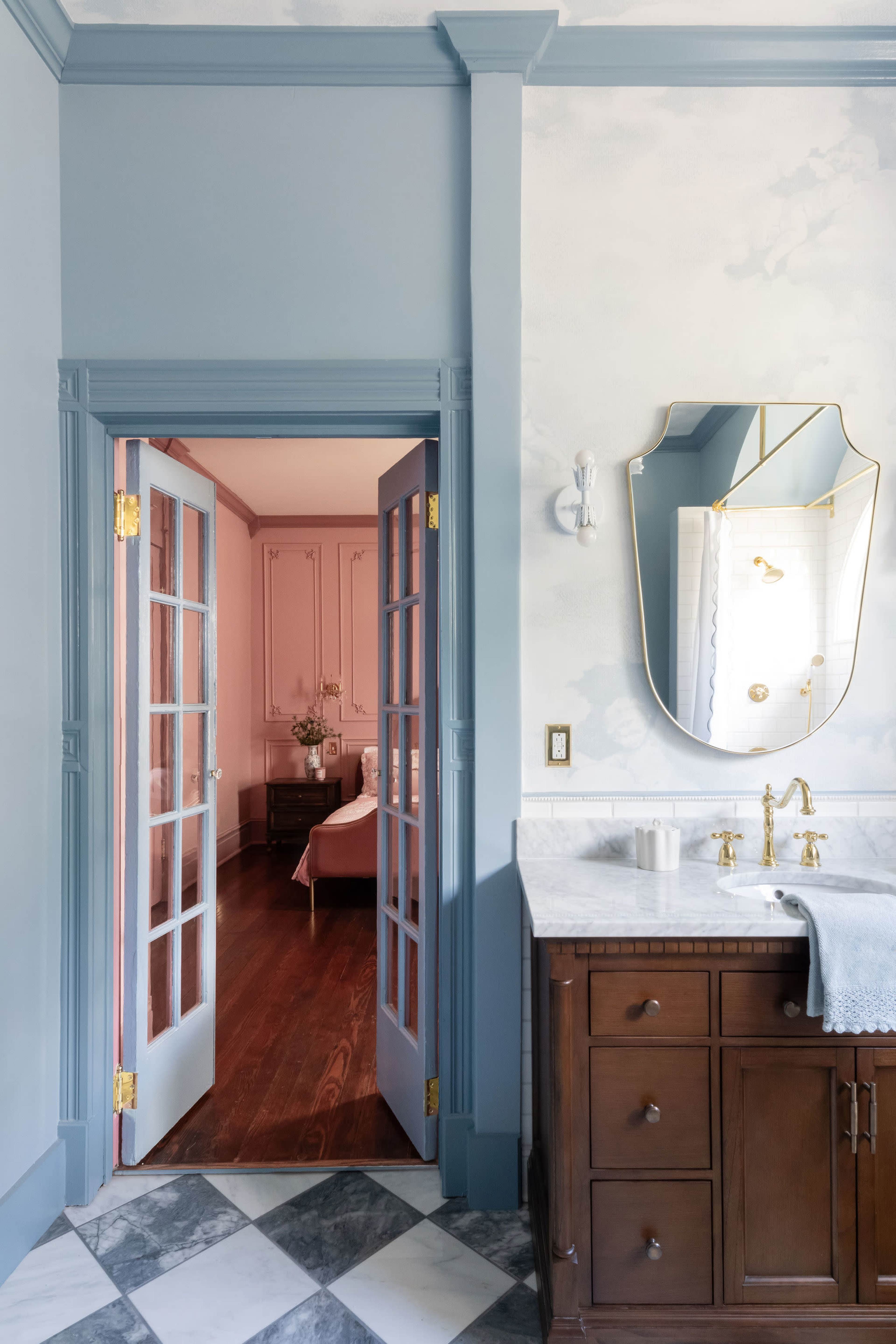 A bathroom features a double vanity with a large mirror, leading to a room adorned in soft pink hues.