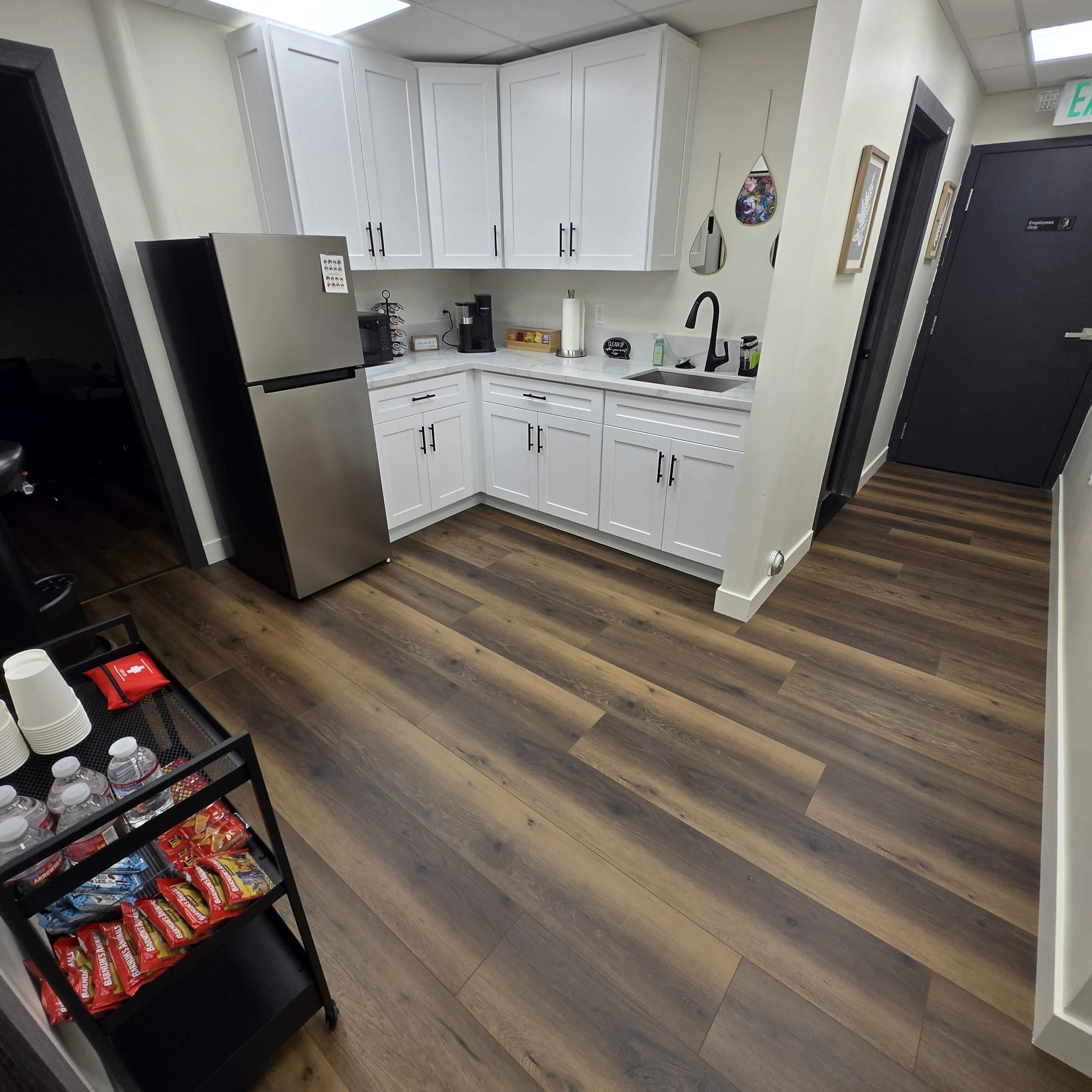 The image shows a modern kitchen area with stainless steel appliances, white cabinetry, and wooden flooring, featuring a beverage station on the left and a hallway leading to another room on the right.