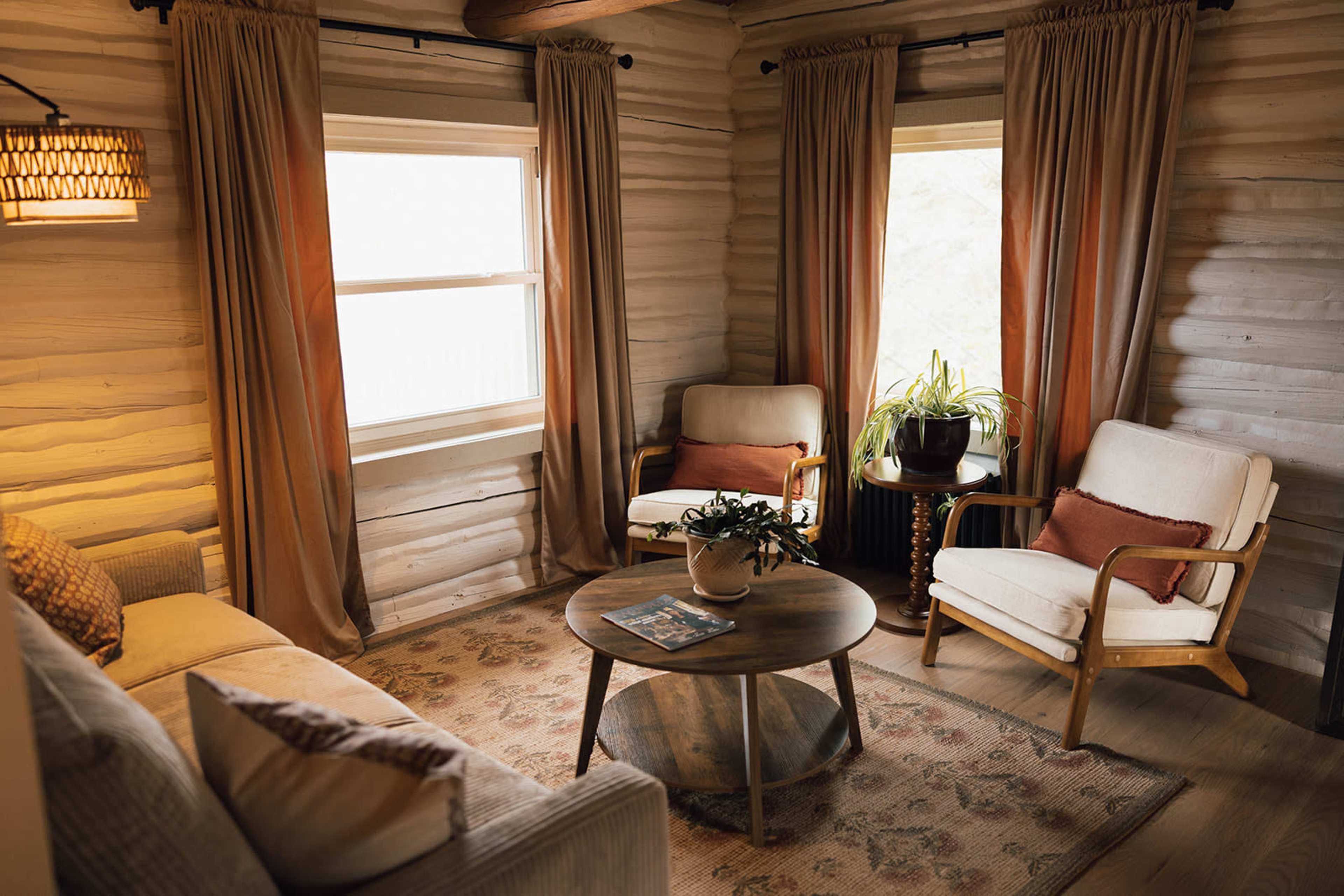 The room features a cozy seating area with two armchairs and a sofa, arranged around a round wooden table, and surrounded by textured walls and soft curtains.