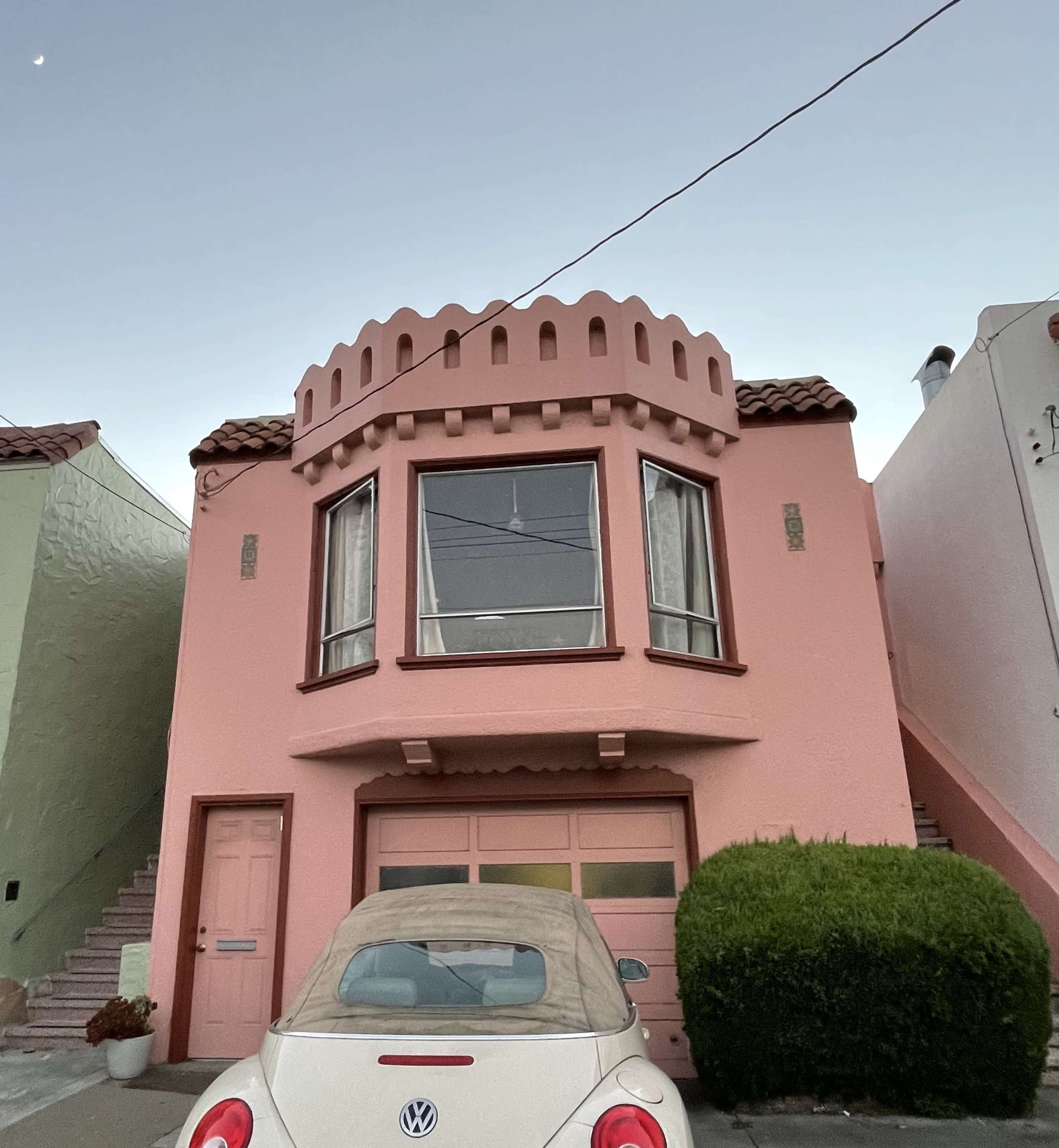 A pink house with a rounded turret and a large window is located above a car parked in front.