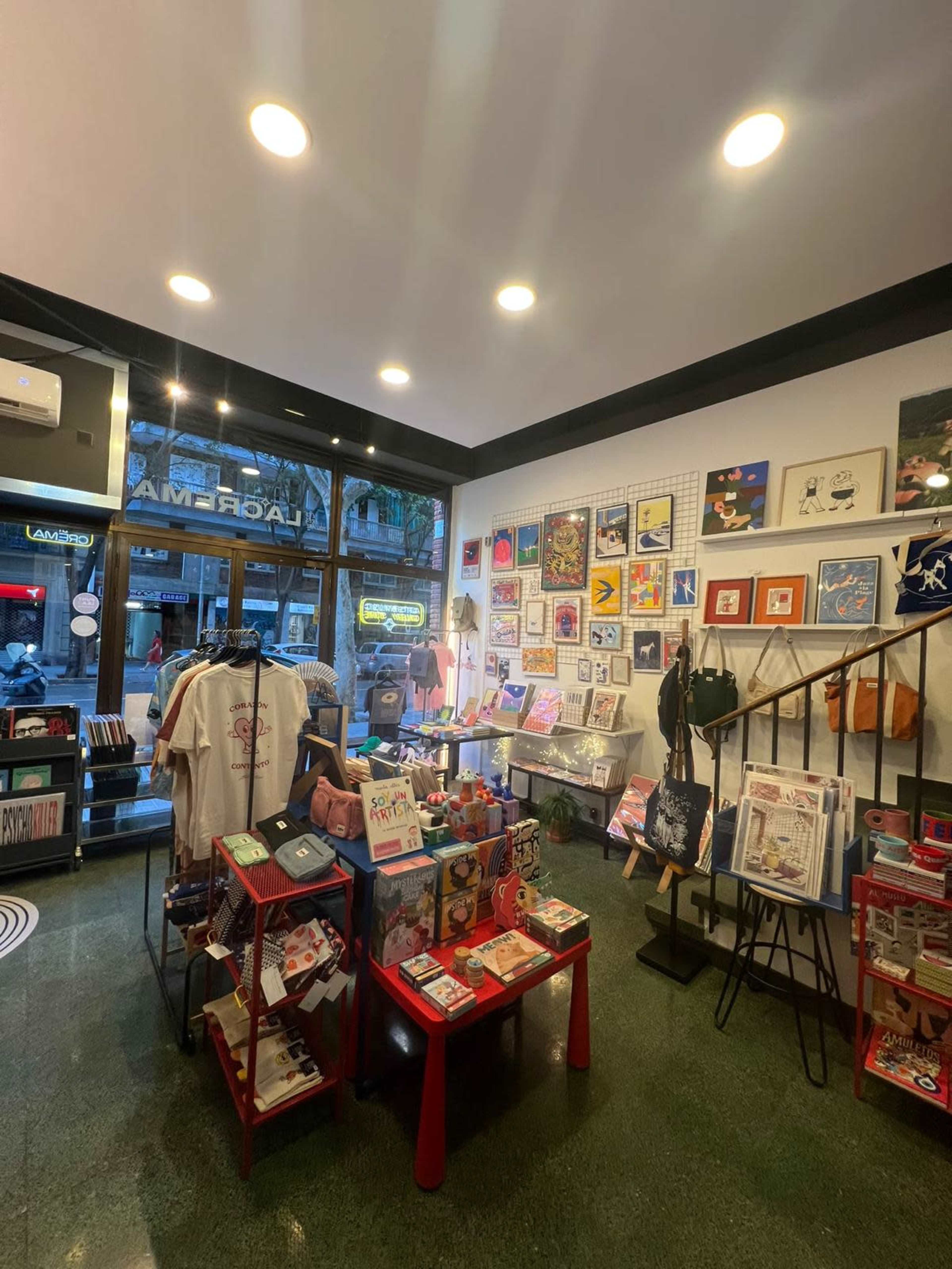 The image shows a vibrant retail space filled with colorful merchandise, including clothing, art prints, and accessories, arranged around tables and shelves.